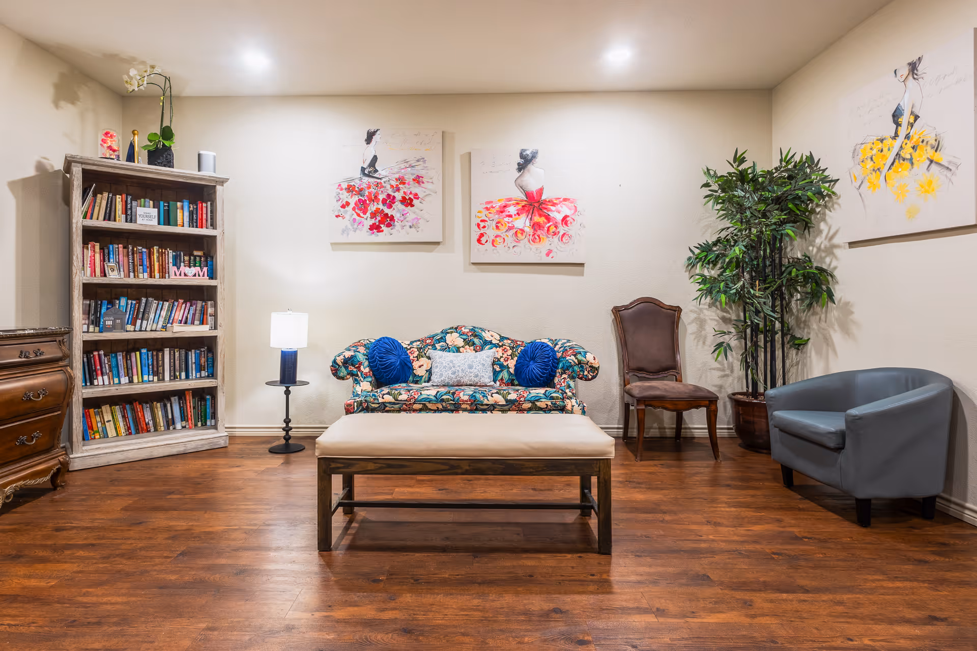 A cozy living room area with a floral-patterned loveseat adorned with blue and white pillows, a beige cushioned bench in front, a wooden bookshelf filled with books on the left, a small round side table with a lamp, a wooden chair, a gray armchair, a tall potted plant, and three colorful paintings of women in dresses on the walls.