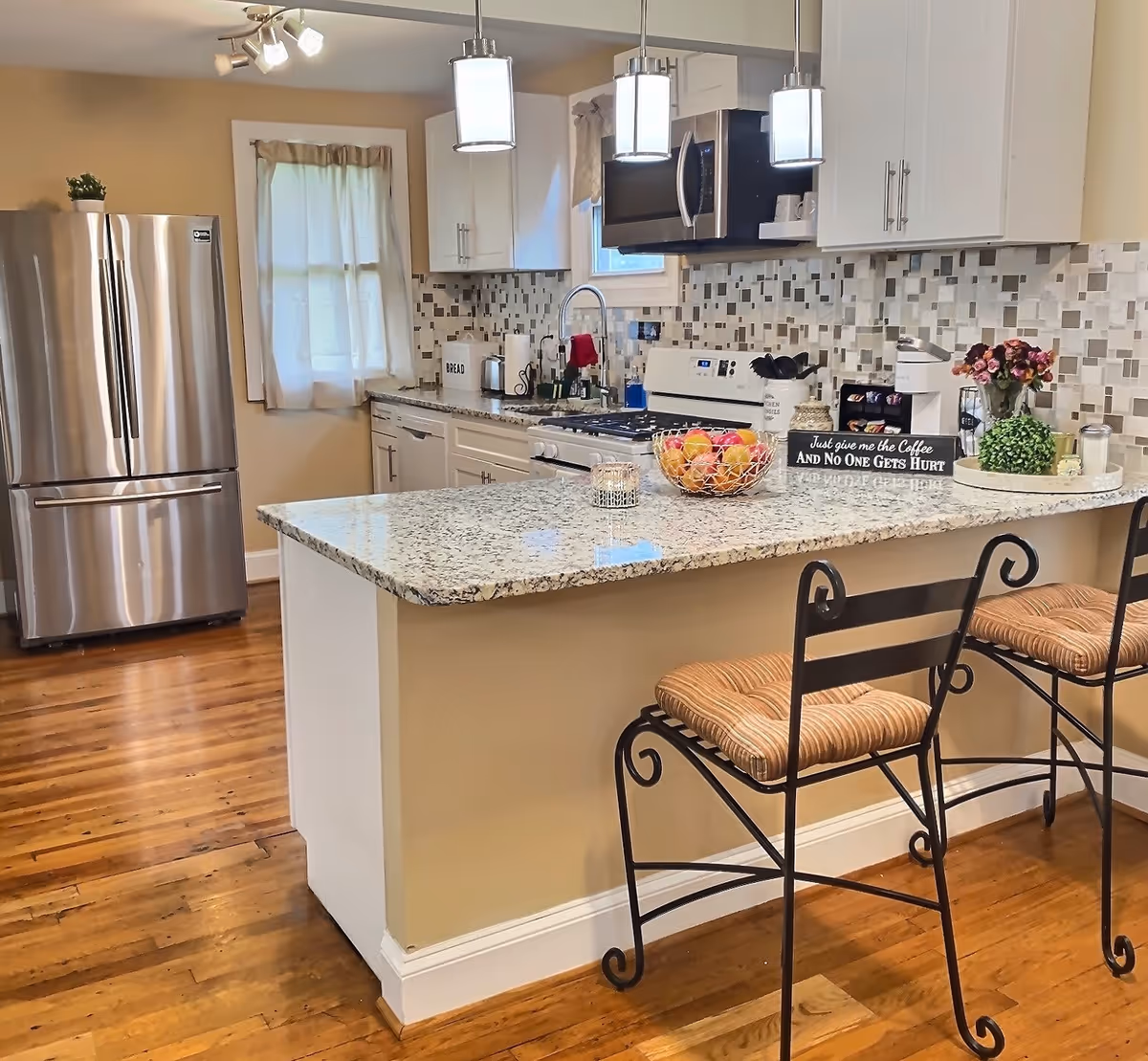 Bright modern kitchen with a granite island and two bar stools, stainless steel refrigerator, stove, and mosaic tile backsplash.