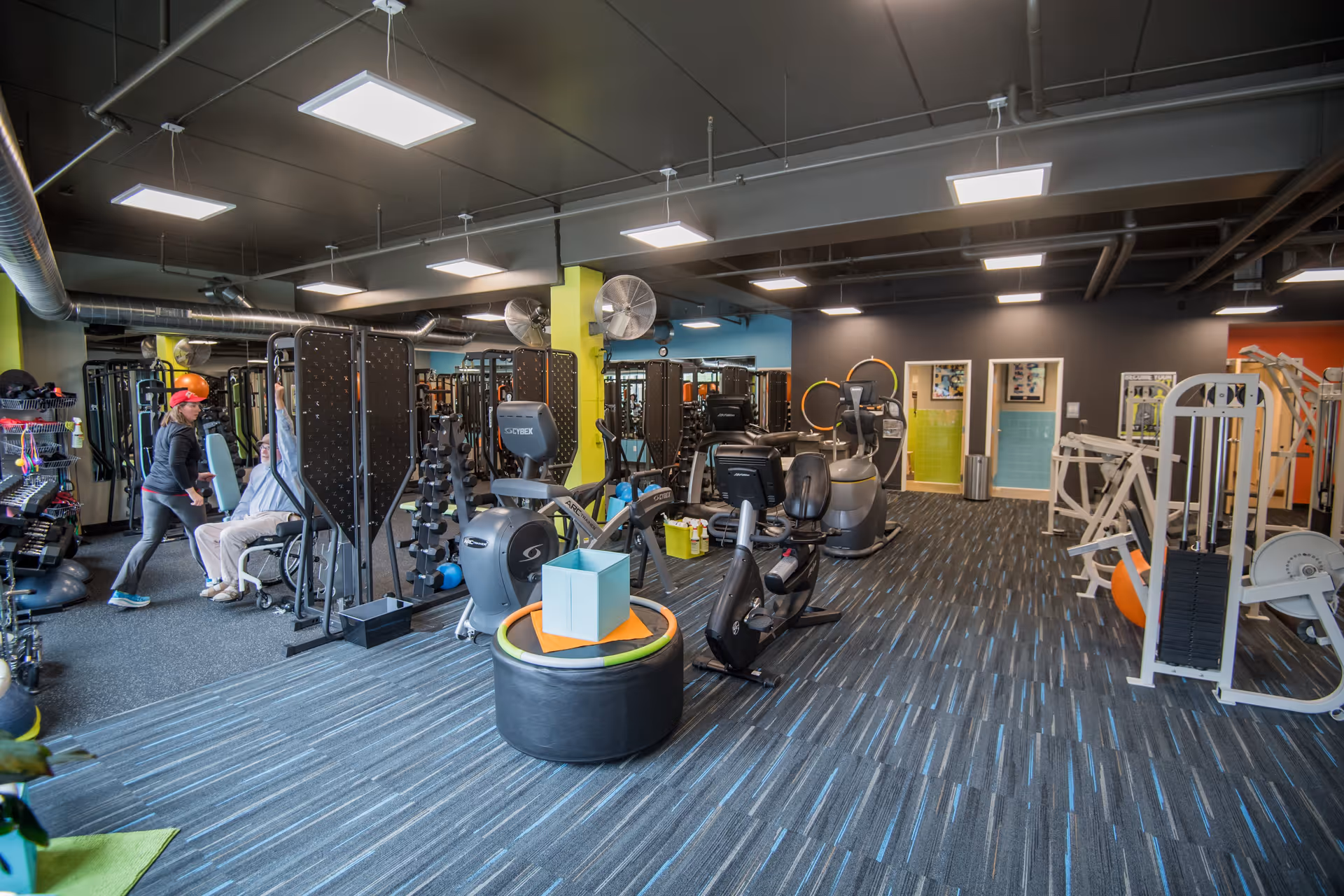 Interior view of a fitness center with exercise equipment including stationary bikes, weight machines, and free weights. Two people are seen exercising on the left side of the room. The room has a modern design with a dark ceiling, bright lighting, and colorful walls.