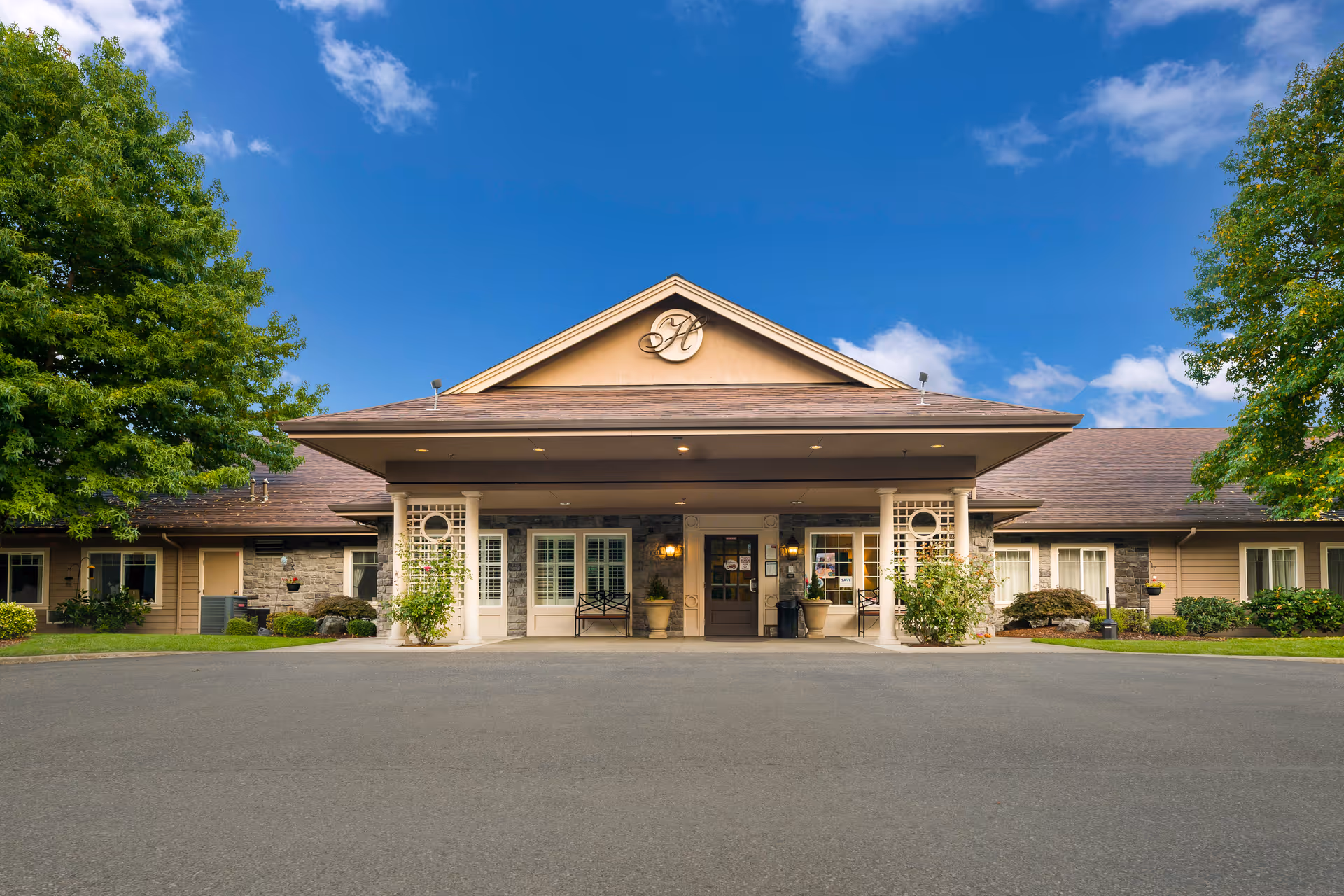 Front exterior view of The Hampton senior living facility building with a covered entrance, surrounded by trees and greenery under a blue sky with some clouds.