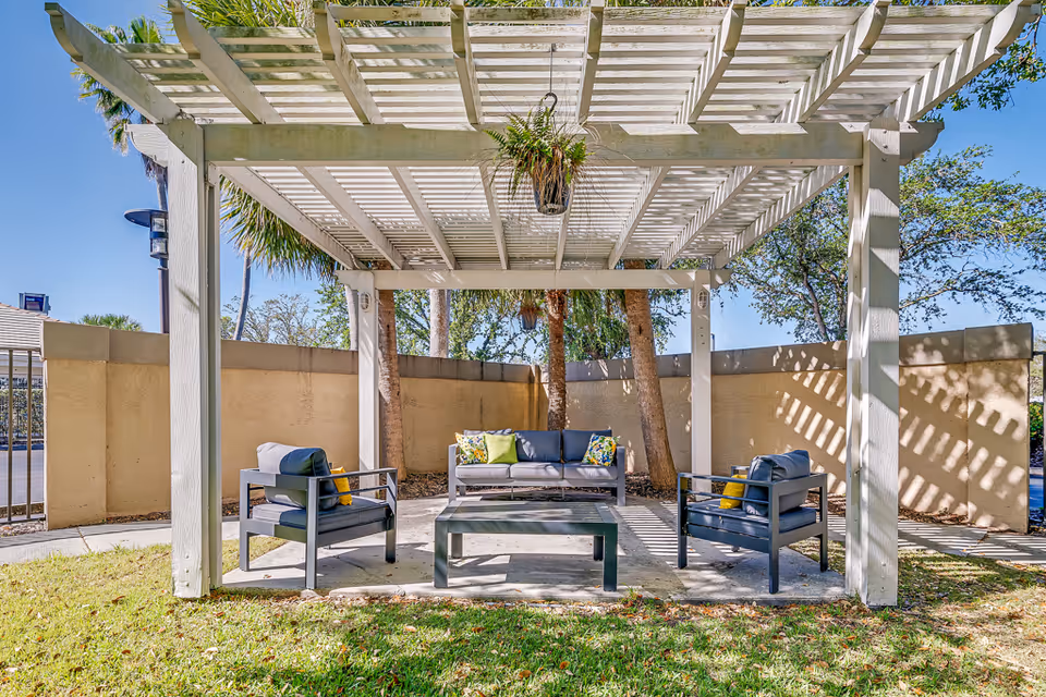 Outdoor seating area under a white pergola with a hanging plant above. The area includes a gray cushioned sofa with colorful pillows and two matching armchairs surrounding a rectangular coffee table. The space is enclosed by a beige wall with trees and a clear blue sky in the background.