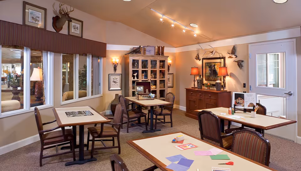 A cozy common room with several tables and chairs arranged for activities and dining. The room features a cabinet with decorative items, wall-mounted lamps, framed artwork, and mounted animal heads and birds. There are windows on one side and a door on the other, with a warm, inviting atmosphere.