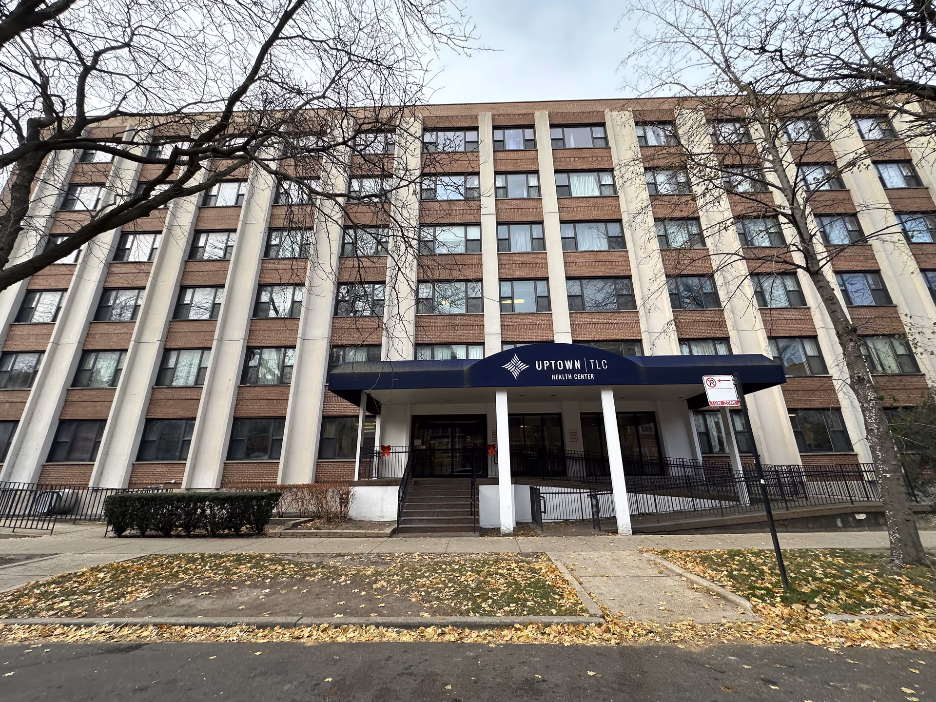 Exterior front view of a multi-story brick and concrete building with a blue awning over the entrance that reads 'Uptown TLC Health Center'. Leafless trees and fallen leaves are visible in the foreground.