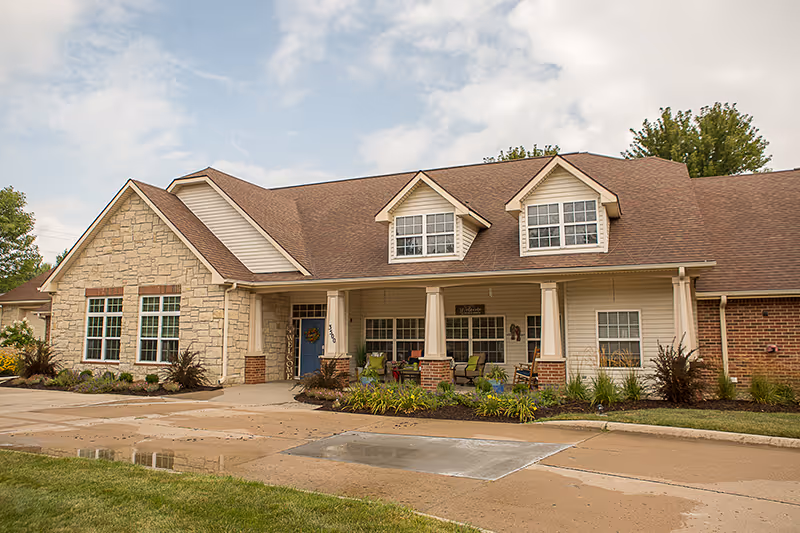 Front exterior of a single-story senior living building with a covered porch, white columns, dormer windows, and landscaped walkway.