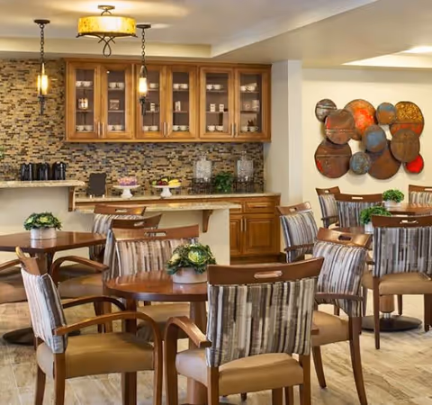 A cozy dining area with several wooden tables and cushioned chairs arranged neatly. Each table has a small green plant centerpiece. The background features a kitchen counter with a mosaic tile backsplash, wooden cabinets with glass doors displaying dishes, and hanging pendant lights. A colorful circular metal wall art piece is mounted on the wall.