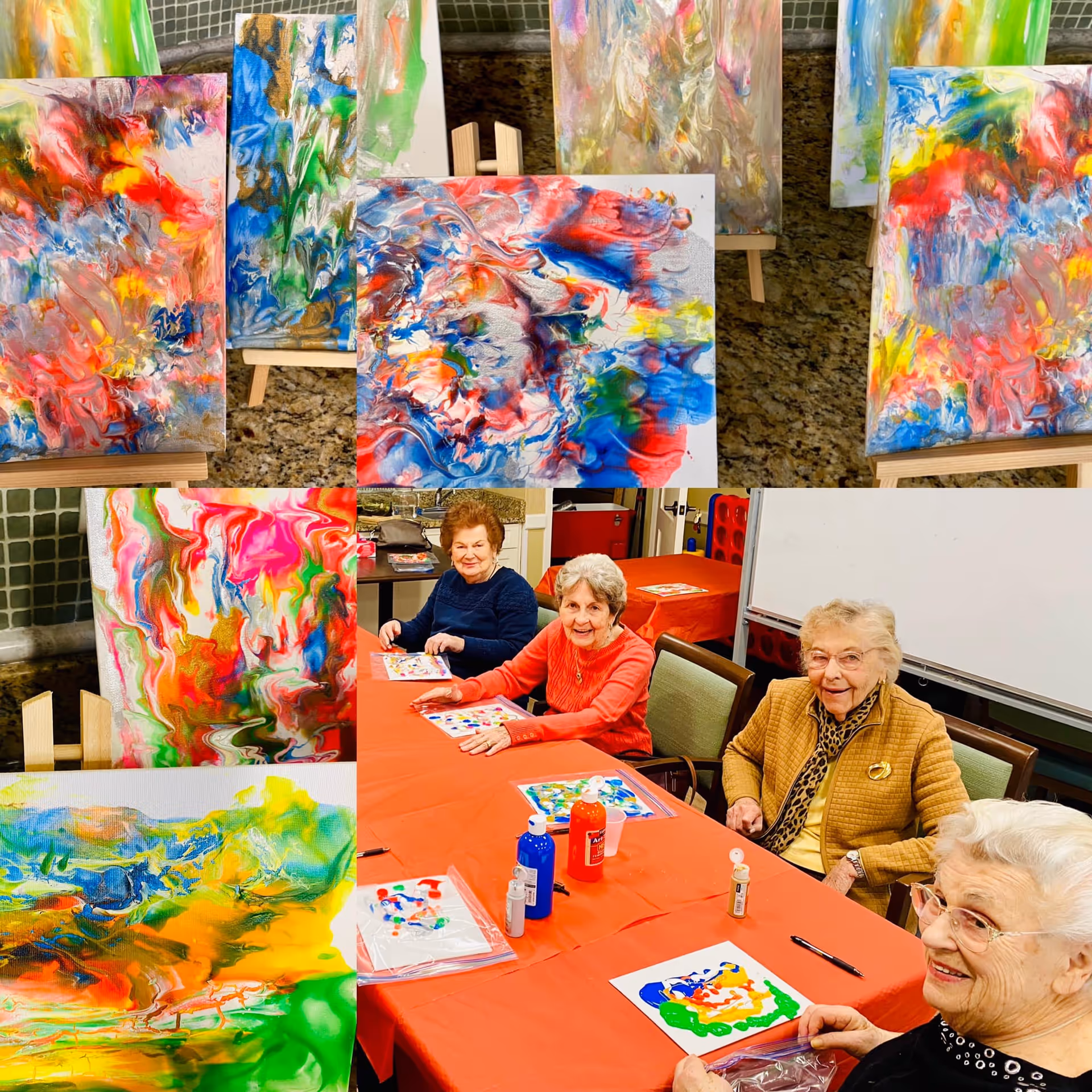 A group of four elderly women sitting around a table covered with an orange tablecloth, engaging in a painting activity with colorful abstract artwork displayed on easels behind them. Various bottles of paint and painting supplies are on the table.