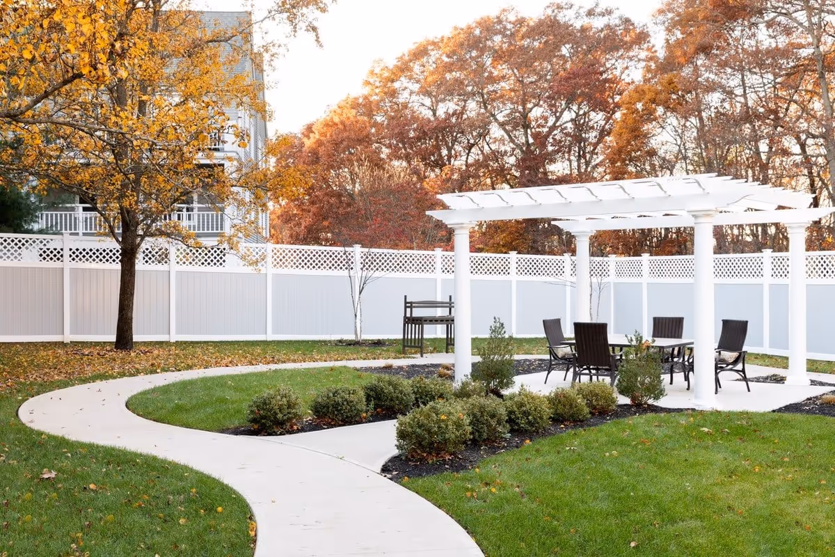 Outdoor garden area with a white pergola covering a table and four chairs. A curved concrete pathway leads to the pergola, surrounded by green grass, small bushes, and trees with autumn-colored leaves. A white fence encloses the area.
