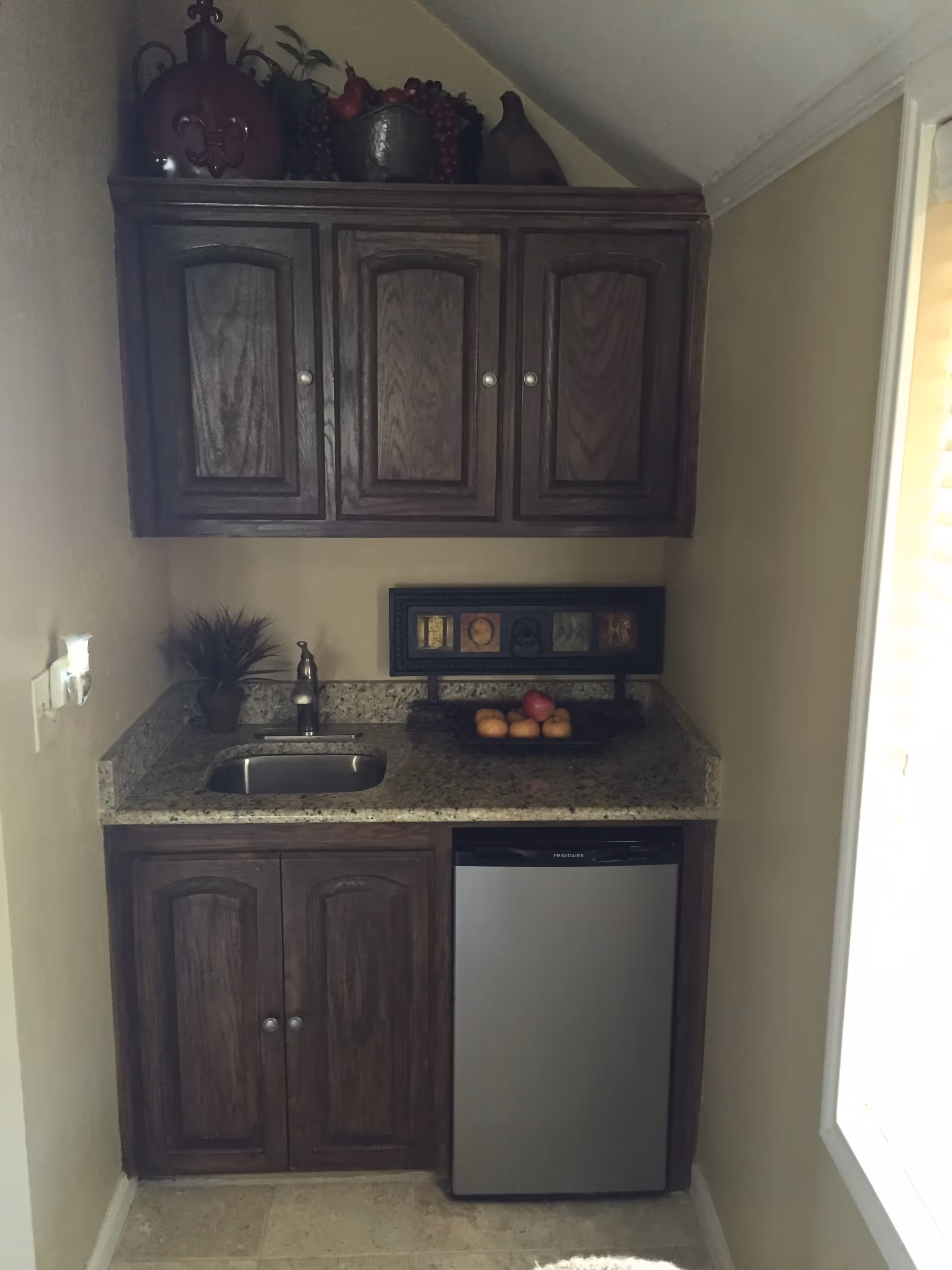 Small kitchenette/wet bar with dark wood upper and lower cabinets, a granite countertop with a small sink, a stainless steel mini fridge, and a decorative bowl of fruit.