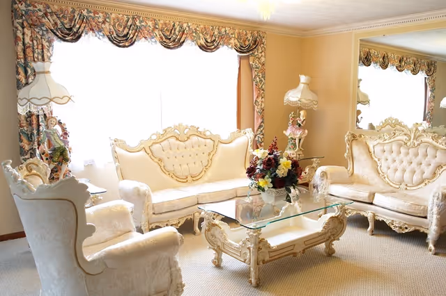 A bright living room with ornate, cream-colored vintage furniture including a sofa, loveseat, and armchair. The room features a glass coffee table with a floral centerpiece, decorative lamps with figurines, floral curtains, and a large mirror on one wall reflecting the furniture.