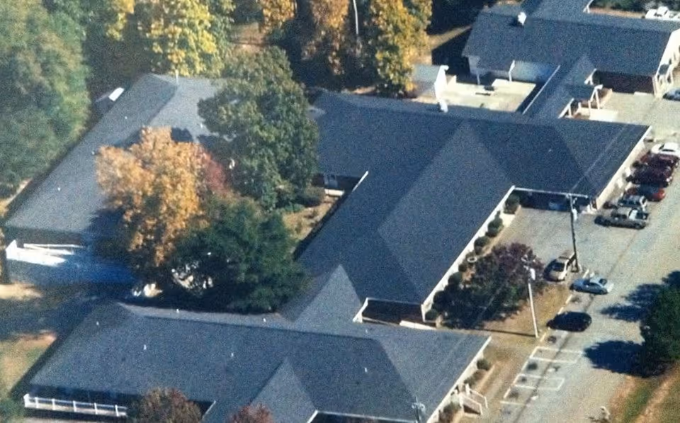 Aerial view of a single-story senior living facility complex with connected dark-roofed buildings, parking lot, and surrounding trees.