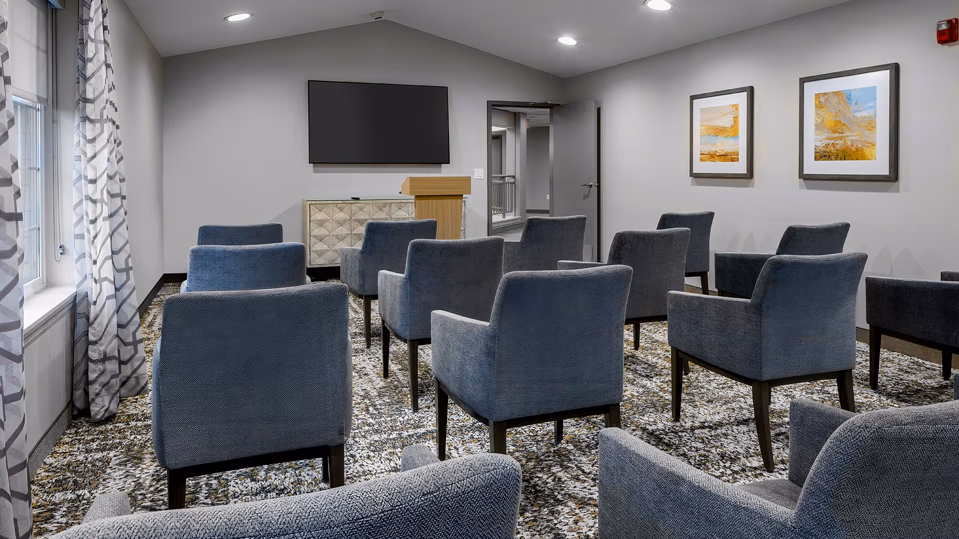 A small conference or presentation room with rows of blue upholstered chairs facing a wall-mounted flat screen TV and a wooden podium. The room has patterned carpet, light gray walls with two framed abstract paintings, and windows with patterned curtains on the left side.