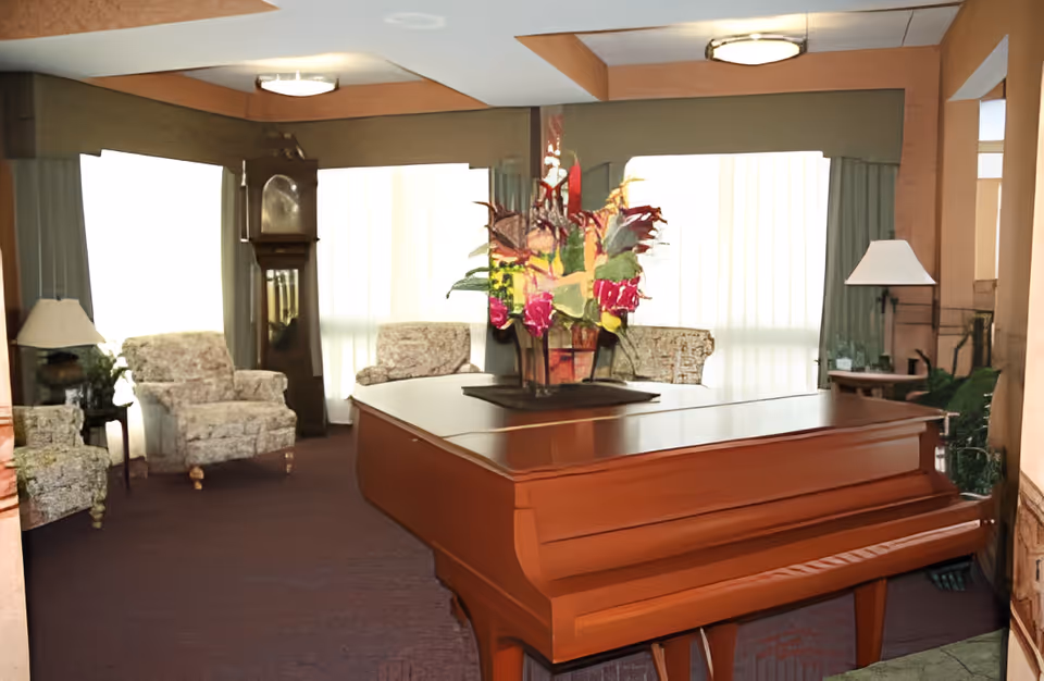 A cozy living room area with a grand piano in the foreground topped with a colorful floral arrangement. In the background, there are several upholstered armchairs, a tall grandfather clock, and large windows with sheer curtains allowing natural light to fill the room. The space has warm wood accents and soft lighting from ceiling fixtures and table lamps.