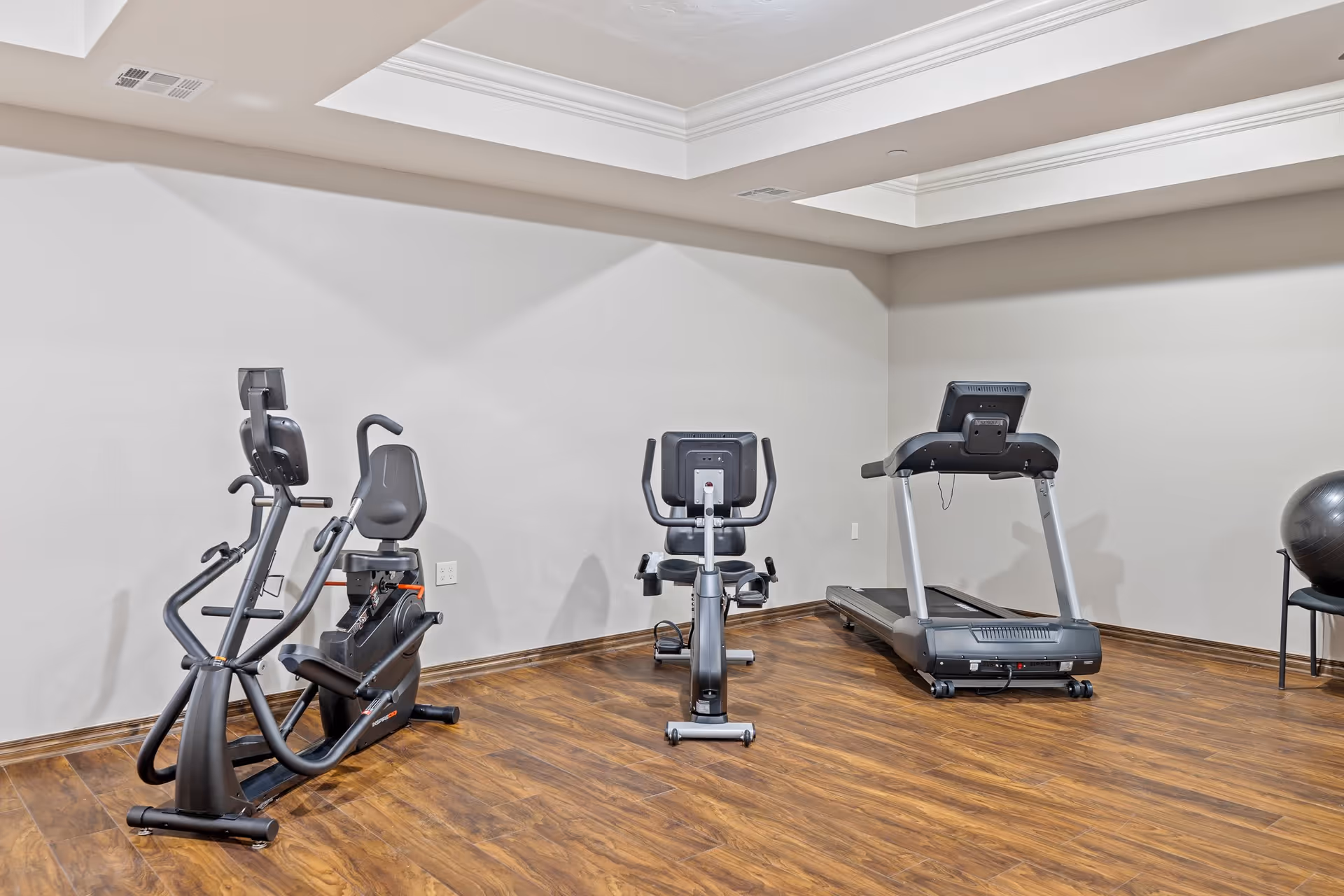 A small fitness room with wooden flooring and light gray walls, featuring two recumbent exercise bikes and a treadmill. There is also a black exercise ball on a chair in the corner.