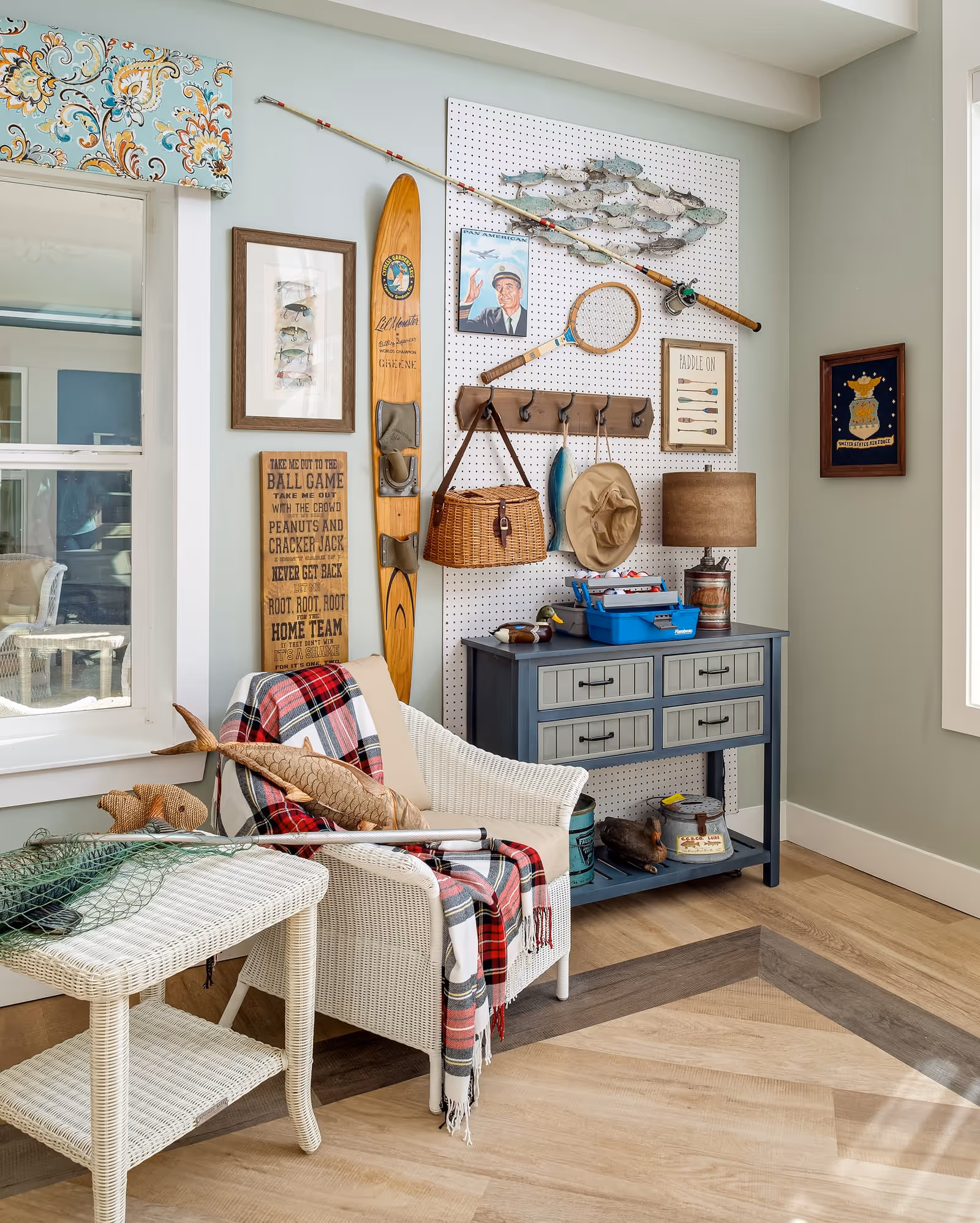 A cozy corner of a room featuring a white wicker chair draped with a red and black plaid blanket and a fish-shaped pillow. Next to the chair is a white wicker side table with a fishing net on top. On the wall behind is a pegboard with fishing-themed decor including a wooden water ski, fishing rod, framed pictures, a wicker basket, a hat, and a wooden rack with hooks. Below the pegboard is a blue-gray console table with drawers and various decorative items on and underneath it. The room has light-colored walls and wood flooring with a decorative inlay.
