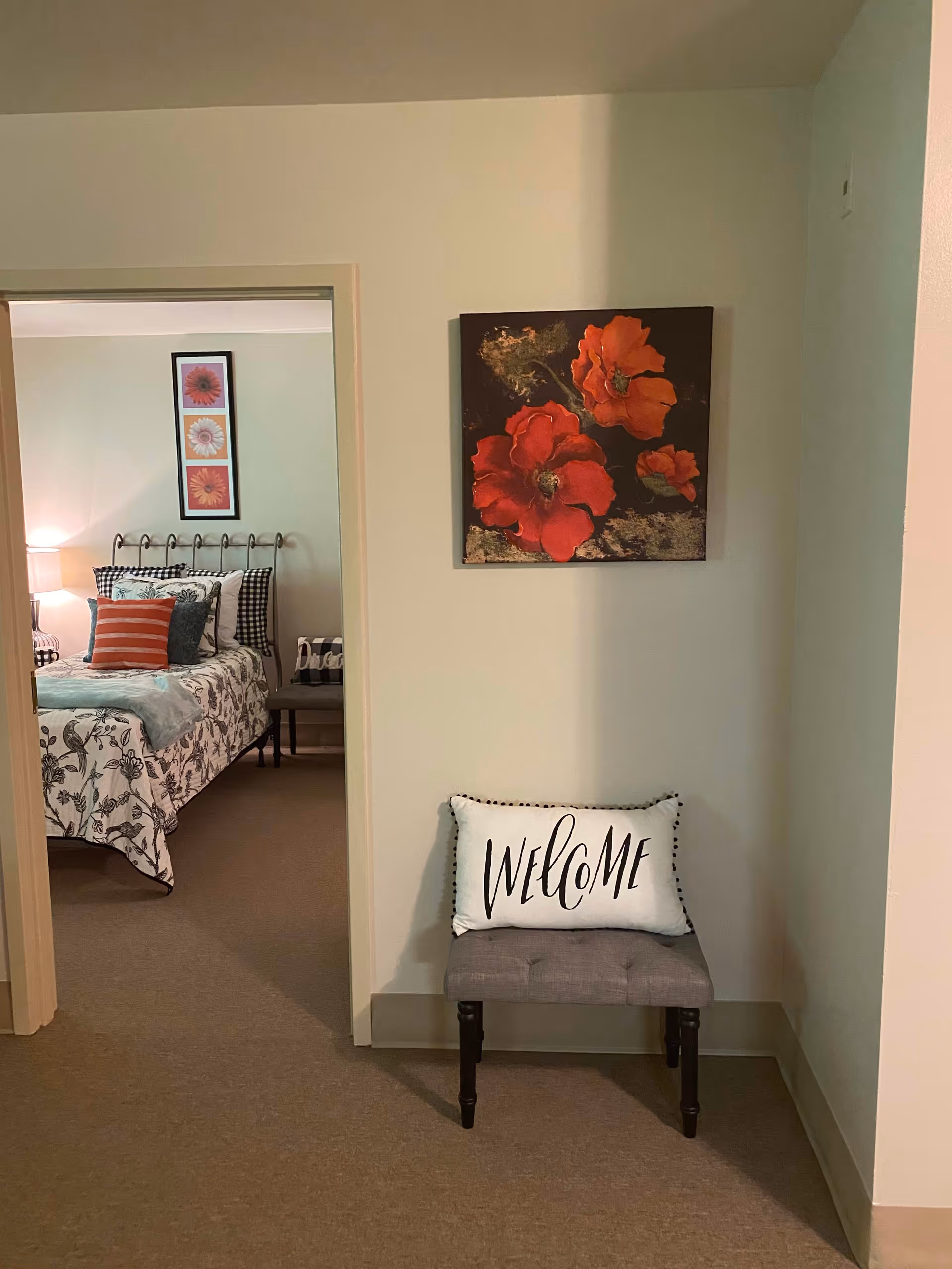 View of a hallway leading to a bedroom in a senior living facility. The hallway has a small cushioned bench with a white pillow that says 'WELCOME'. Above the bench is a wall painting featuring large red flowers. The bedroom visible through the doorway has a bed with floral bedding, multiple pillows including a red striped one, a bedside lamp, and framed floral artwork on the wall.