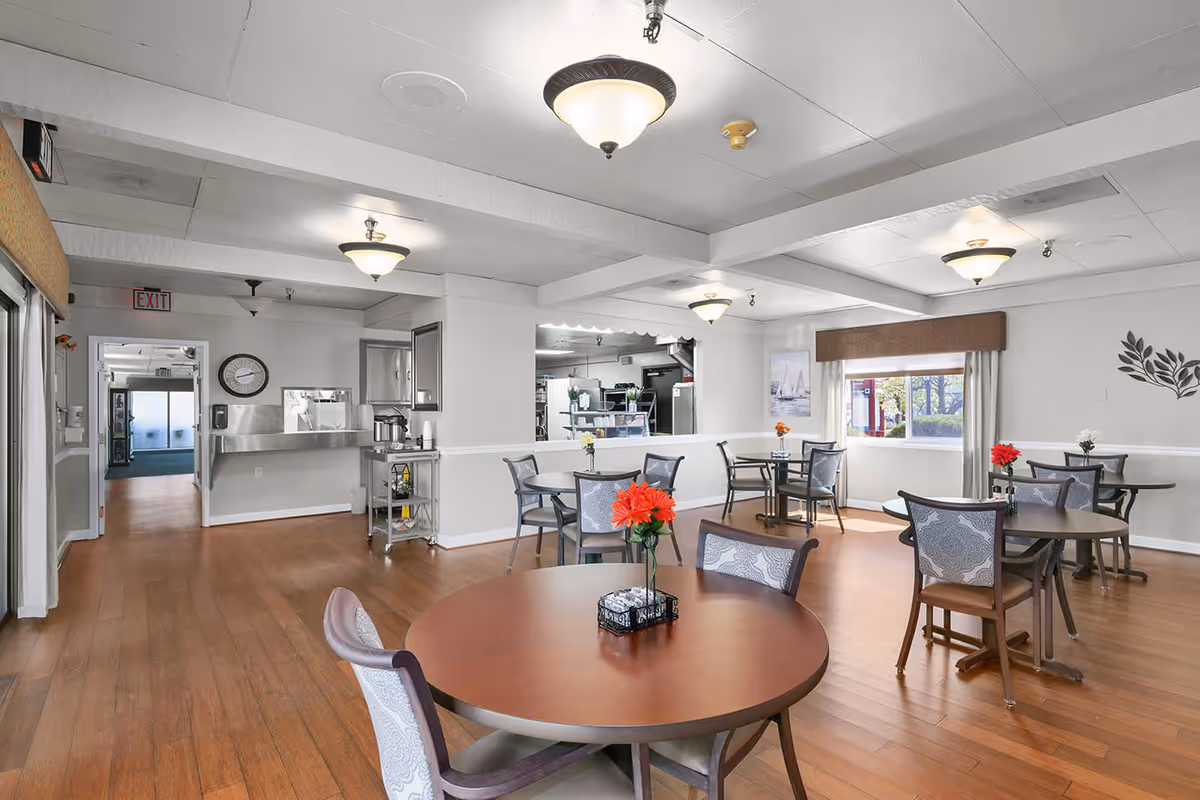 Communal dining room with round tables and chairs, wood floors, ceiling lights, and a serving area in the back.