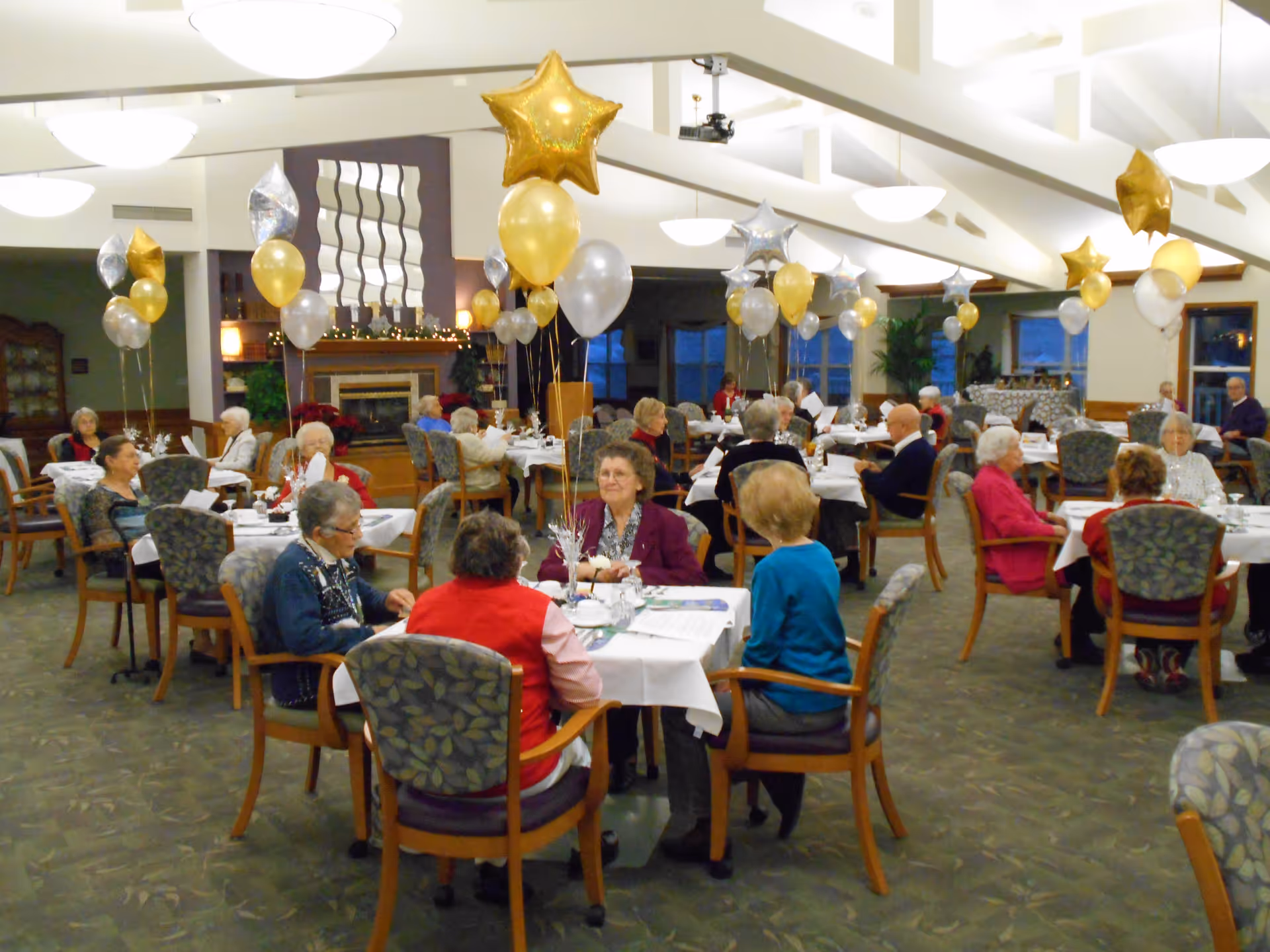 A large dining room decorated with gold and silver star-shaped and round balloons. Several elderly people are seated at tables covered with white tablecloths, engaging in conversation. The room has a high ceiling with hanging lights and a fireplace in the background.