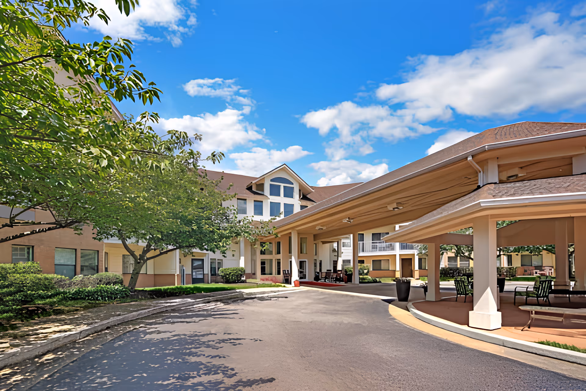 Front entrance of a senior living facility with a covered porte-cochere, driveway, and landscaped trees under a blue sky.