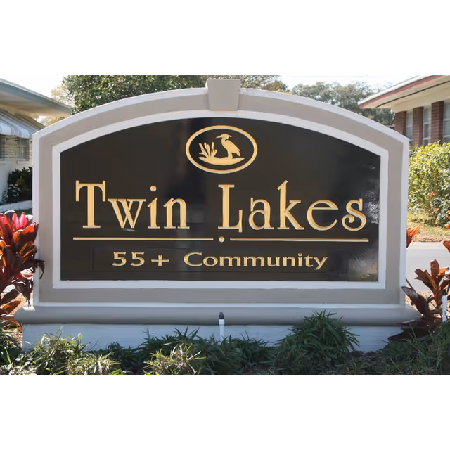 A large black and white sign with gold lettering that reads 'Twin Lakes 55+ Community' with a small bird and reeds emblem above the text, surrounded by greenery and plants.
