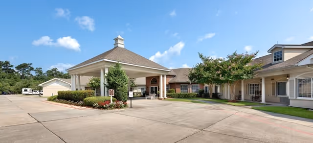 Exterior view of Brookdale Mandeville senior living facility showing a covered entrance with columns, a driveway, landscaped bushes, trees, and a clear blue sky with a few clouds.
