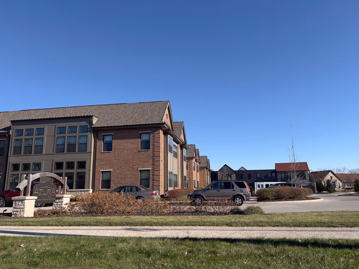Front view of a multi-story brick senior living building with a landscaped driveway, parked cars, and a large sign in front under a clear blue sky.