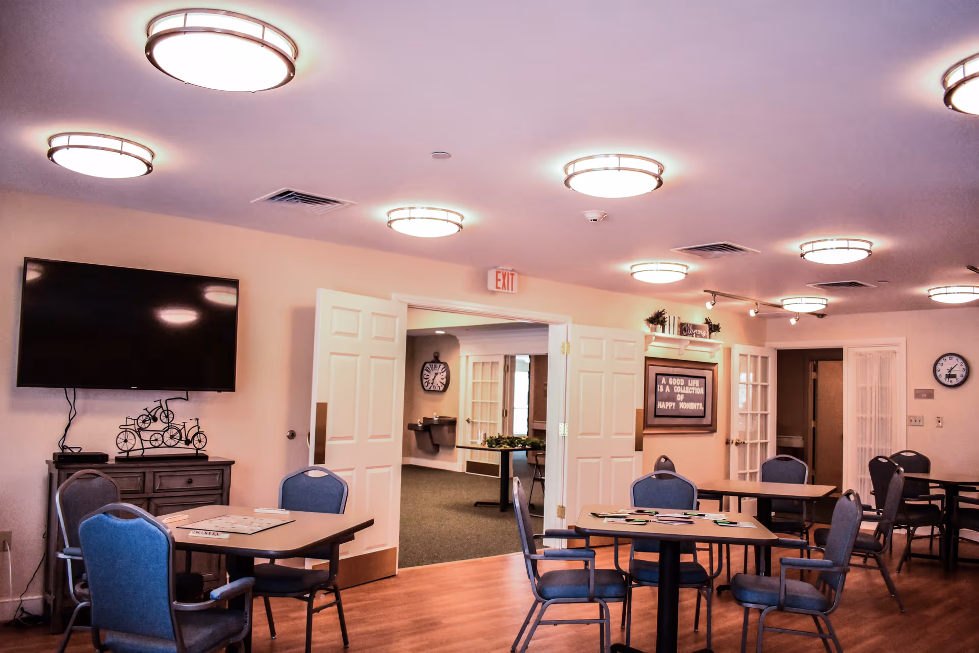 A well-lit common area with multiple tables and chairs arranged for group activities. A flat-screen TV is mounted on the wall above a wooden cabinet with decorative items. Double doors open to another room with a water fountain and wall clock. A framed sign on the wall reads 'A GOOD LIFE IS A COLLECTION OF HAPPY MOMENTS.' The room has wooden flooring and ceiling lights.