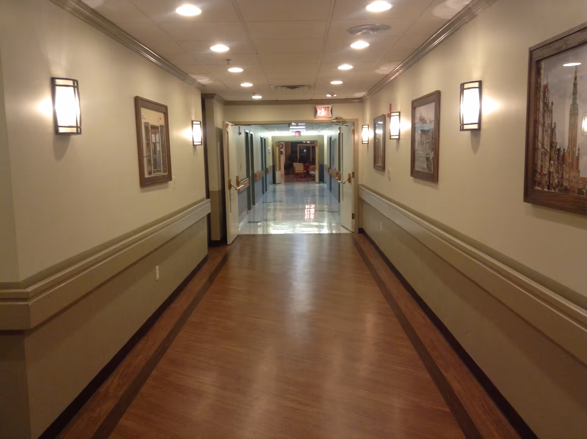 A well-lit indoor hallway with wooden flooring and beige walls, decorated with framed pictures and wall-mounted lights. The hallway has handrails on both sides and leads to an open double door at the end, revealing another corridor with chairs visible in the distance.