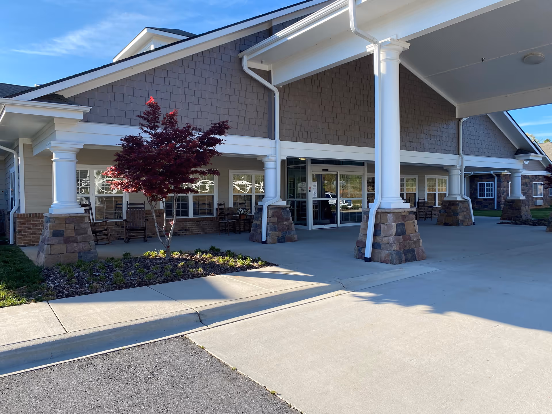 Front exterior view of Grace Village Assisted Living and Memory Care building with a covered entrance supported by white columns with stone bases, a small landscaped area with a red-leafed tree, and several rocking chairs on the porch.