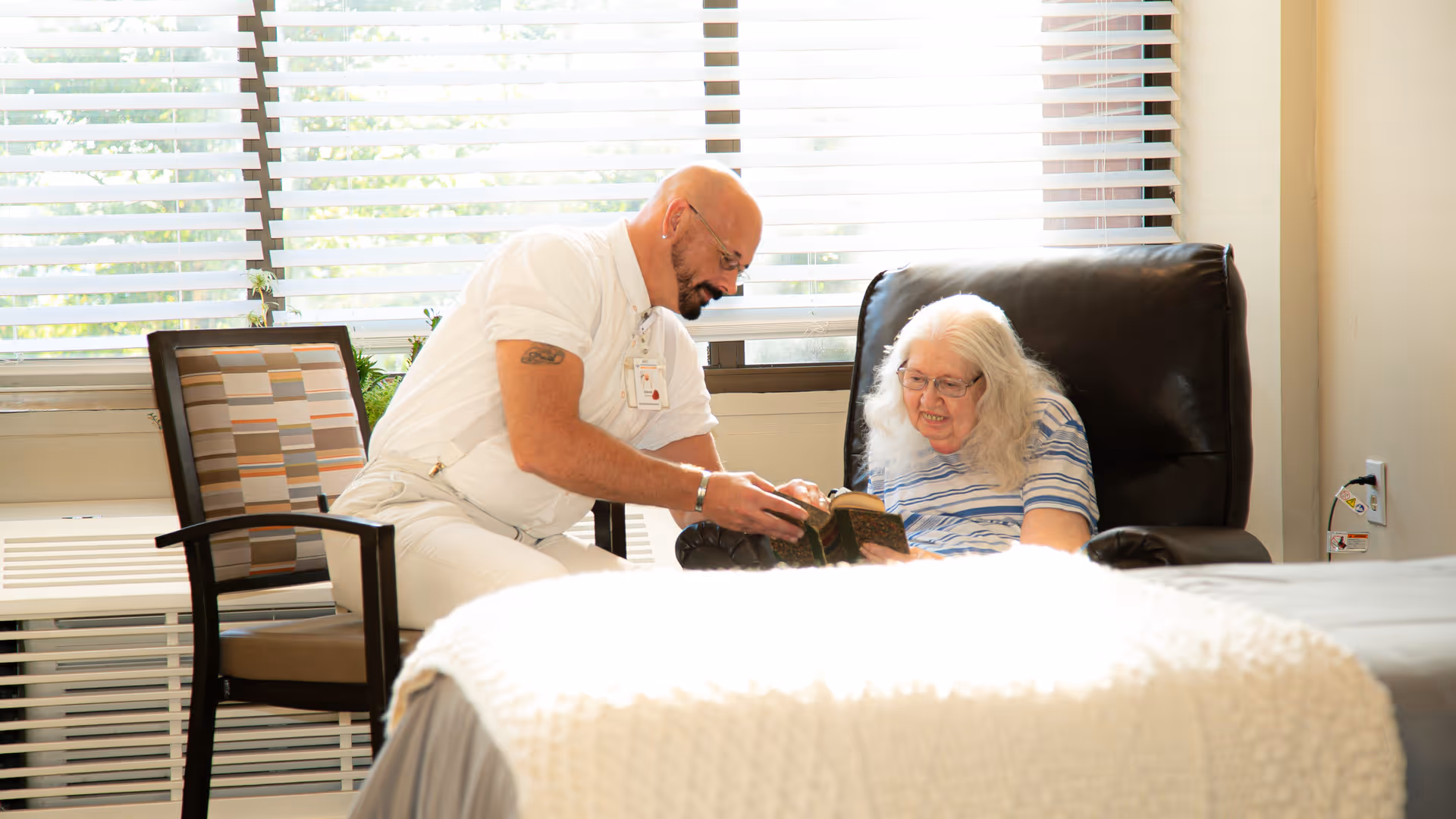A caregiver in white uniform is sitting next to an elderly woman with long white hair who is seated in a black recliner chair. They are looking at a book together in a well-lit room with large windows covered by horizontal blinds. A bed with a white knitted blanket is visible in the foreground.