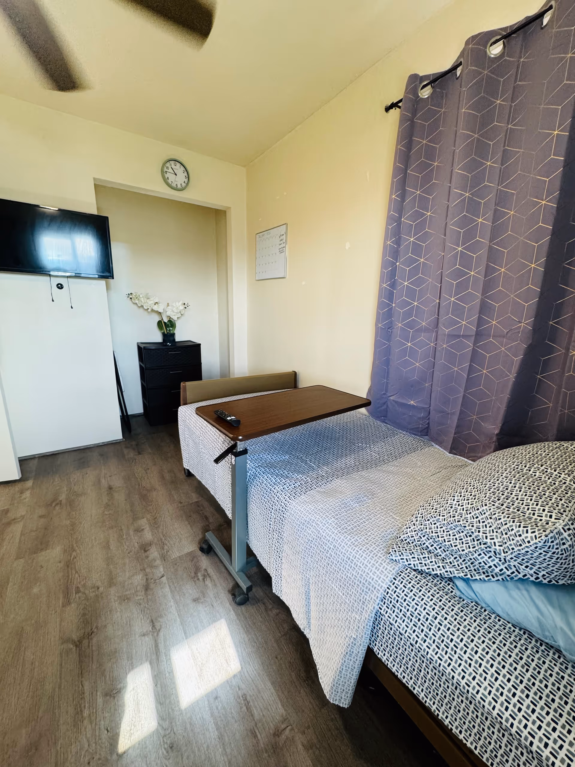 A small bedroom with a single bed covered in a patterned bedspread and pillows. A rolling overbed table with a remote control on it is positioned over the bed. There is a window with purple curtains on the right wall. On the far wall, a flat-screen TV is mounted, a clock hangs above a small black dresser with a vase of white flowers, and a calendar is mounted on the wall. The floor is wood laminate.