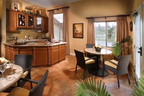 A cozy dining area with a round table surrounded by four cushioned chairs, adjacent to a wooden kitchen counter with cabinets and a sink. The room has large windows with beige curtains letting in natural light, a potted plant near the window, and a framed artwork on the wall.
