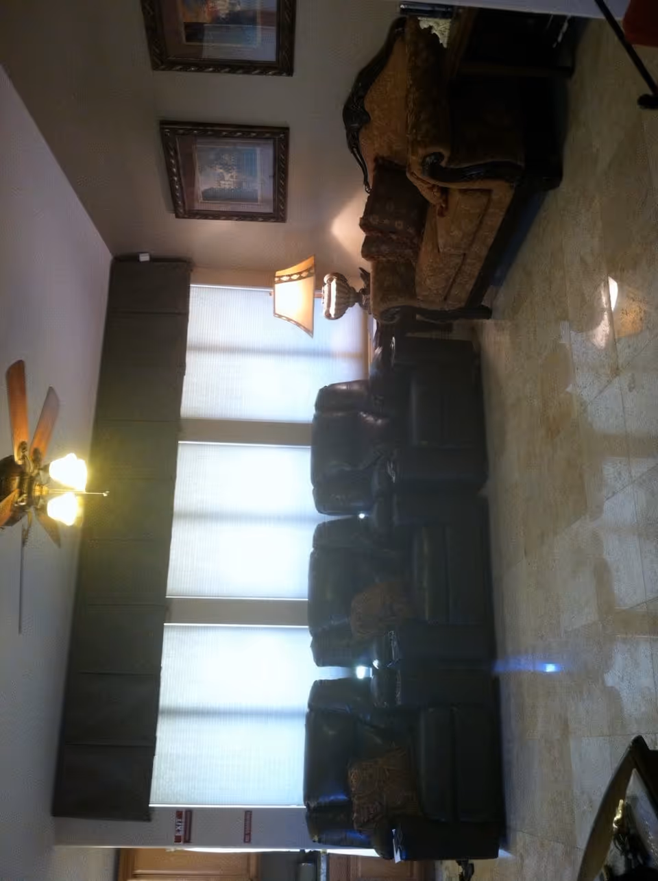 A living room with a ceiling fan and lights, a large window covered with blinds and a dark valance, a table lamp on a side table, a patterned sofa with cushions, and two dark leather recliners with cushions on a tiled floor.