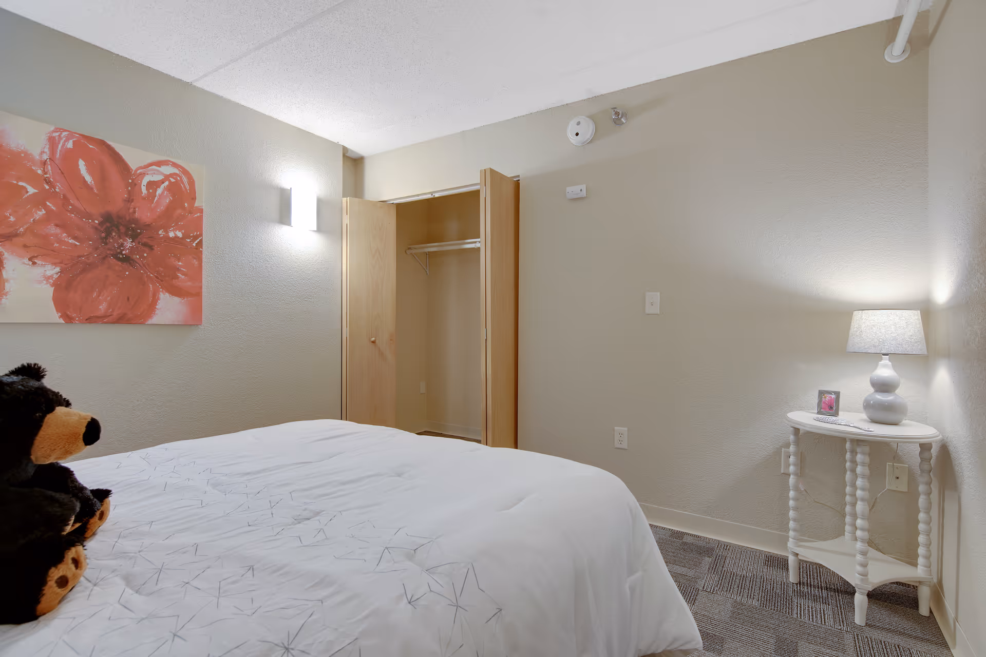 A bedroom with a bed covered in a white comforter featuring a subtle star pattern. A teddy bear is sitting on the bed. On the right side, there is a small white round table with a lamp and a small framed picture. The walls are beige, and there is an open closet with wooden doors. A large painting of a red flower hangs on the left wall.