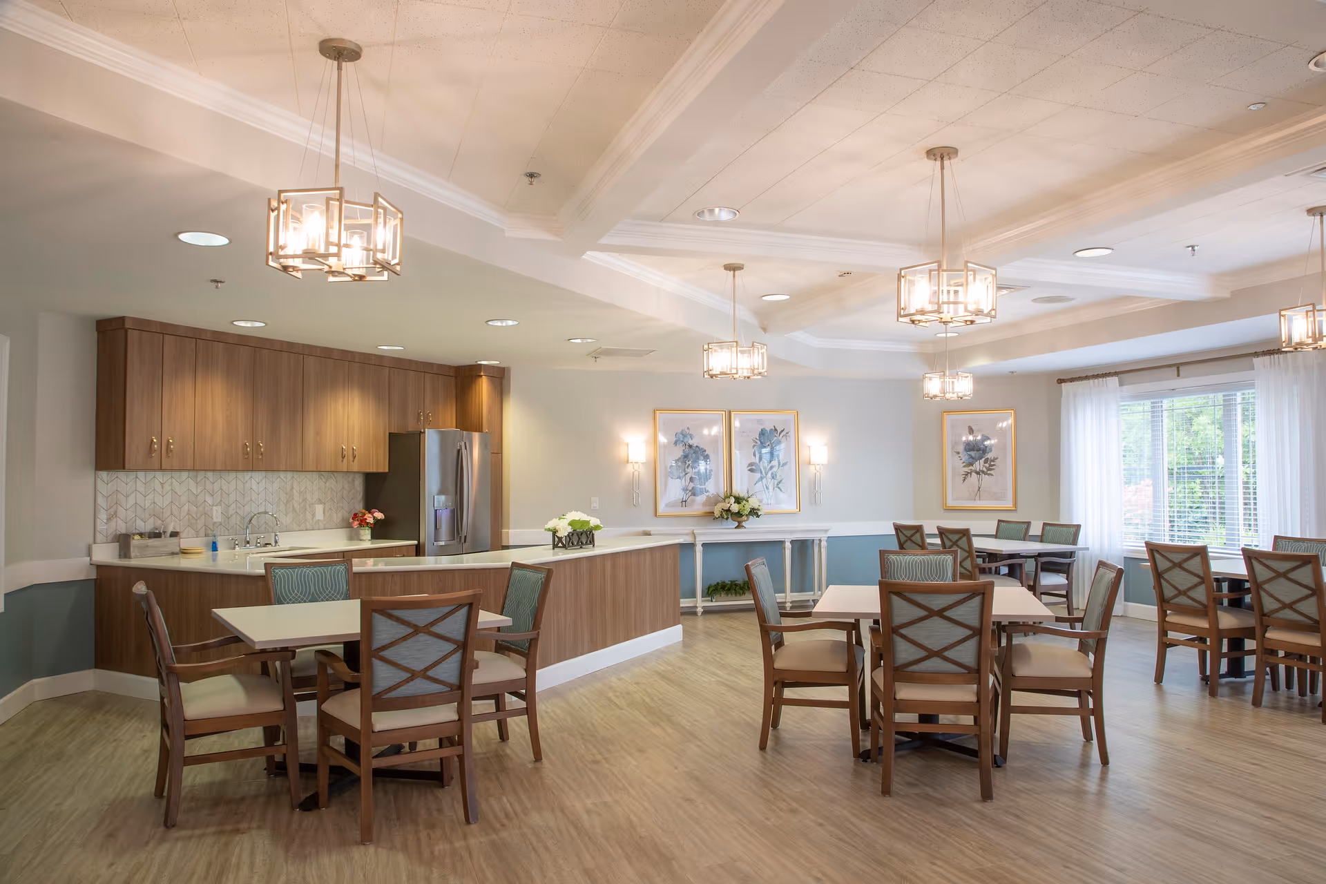 A bright and spacious dining area in a senior living facility with multiple wooden tables and chairs arranged neatly. The room features a kitchen area with wooden cabinets, a stainless steel refrigerator, and a countertop. Modern pendant lights hang from the ceiling, and large windows with white curtains allow natural light to fill the space. The walls are decorated with framed floral artwork and wall sconces.