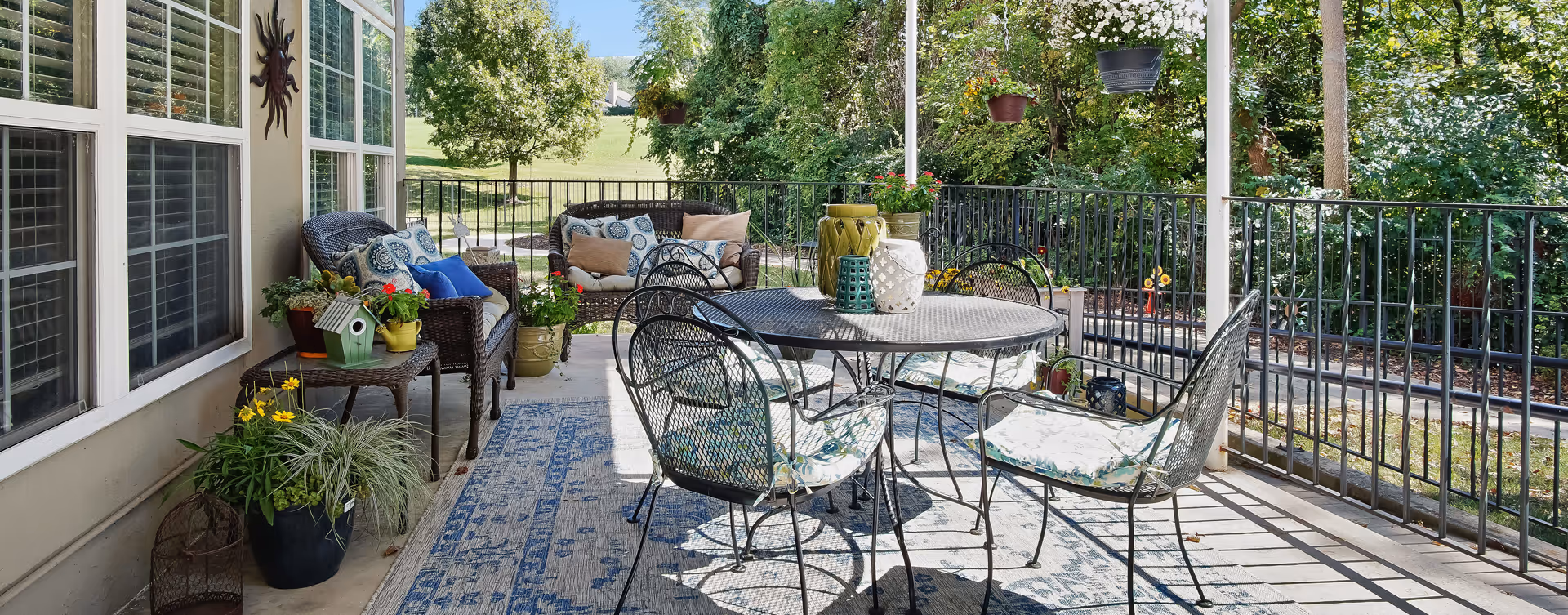 A cozy outdoor patio area with a round metal table and four cushioned chairs. There are two wicker loveseats with decorative pillows, various potted plants, hanging flower pots, and a blue patterned outdoor rug. The patio is surrounded by a black metal railing and overlooks a green lawn and trees.