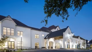 Exterior view of The Blake at Baxter Village senior living facility at dusk, showing a large white building with multiple windows, a covered entrance, and a tree branch in the foreground.
