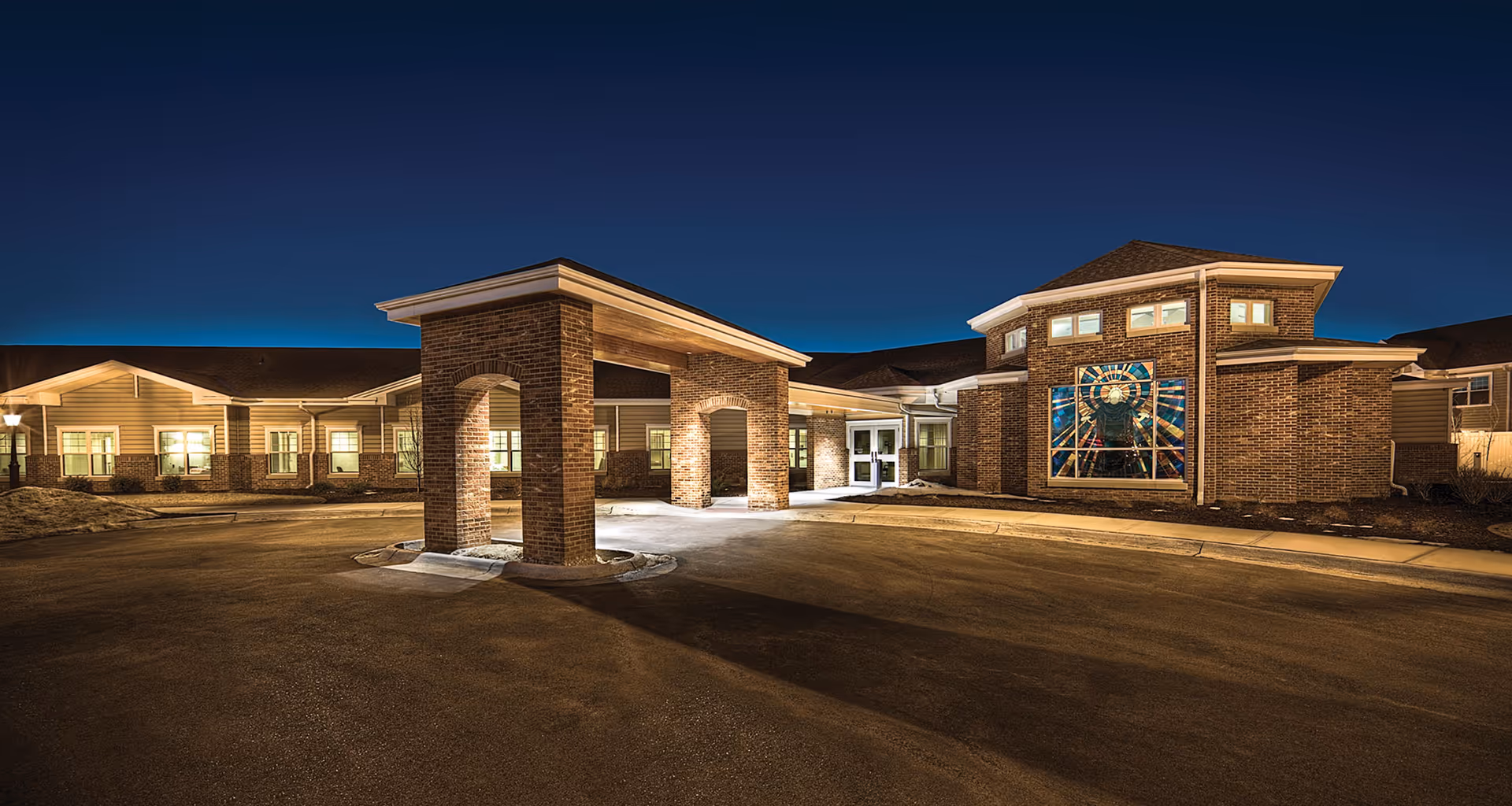 Night view of the front exterior of a senior living facility named Immanuel: The Lighthouse, featuring a brick building with a covered entrance and illuminated windows. A large stained glass window is visible on the right side of the building.