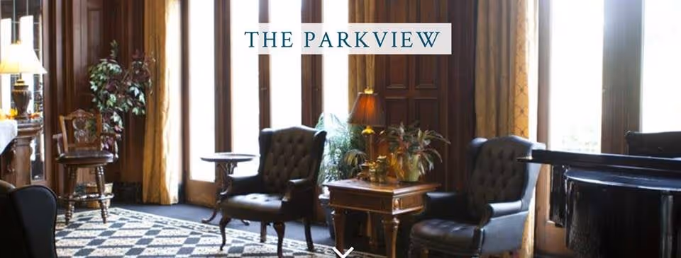 A cozy living room area with two dark leather armchairs, a wooden side table with plants and a lamp, large windows with curtains letting in natural light, and a patterned rug on the floor. The room has wooden paneling and a warm, inviting atmosphere.