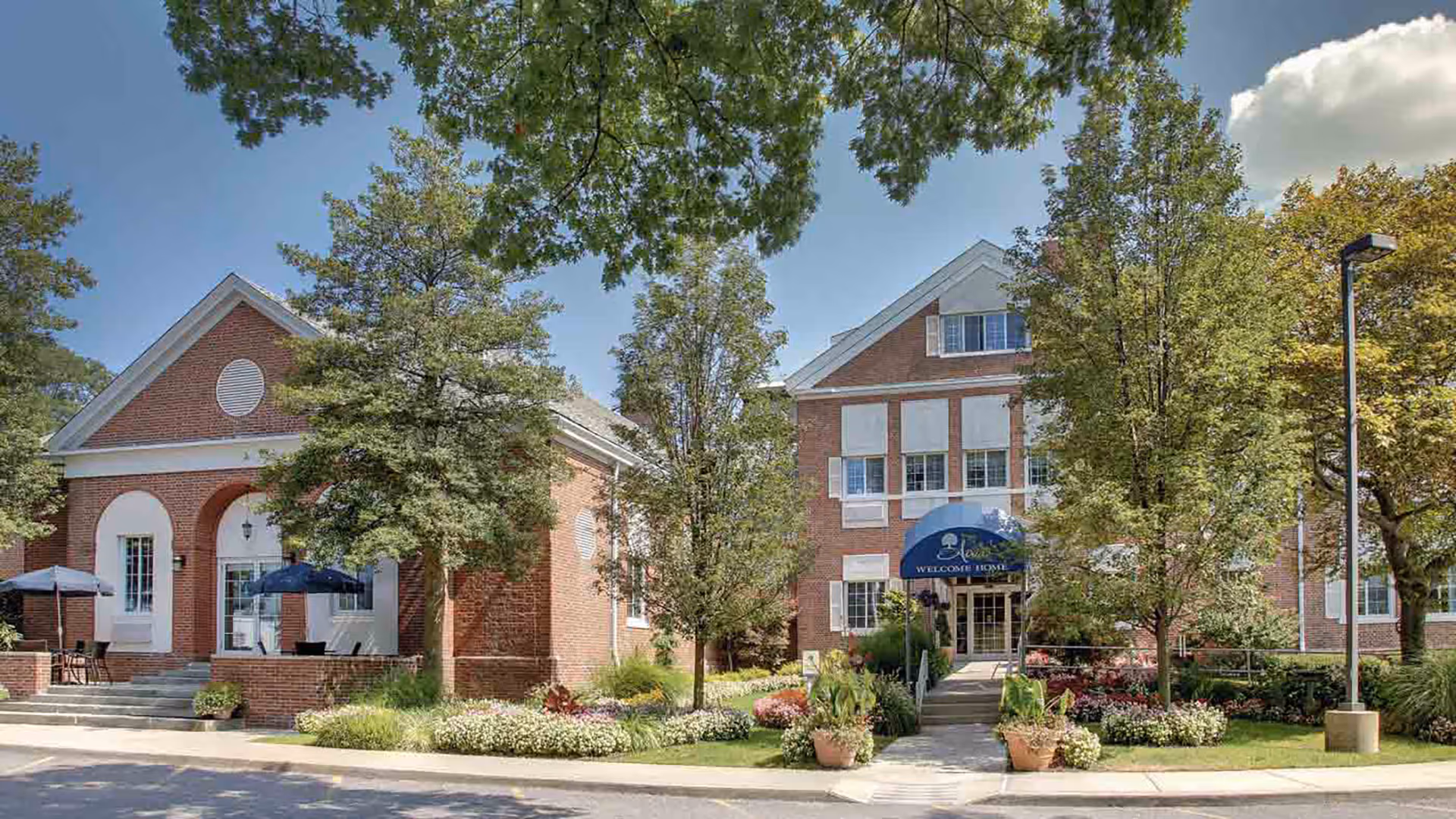 Exterior view of a senior living facility named Atria East Northport, featuring red brick buildings with white trim, a blue awning over the entrance that reads 'Welcome Home,' surrounded by well-maintained landscaping with trees, shrubs, and flowers under a clear blue sky.