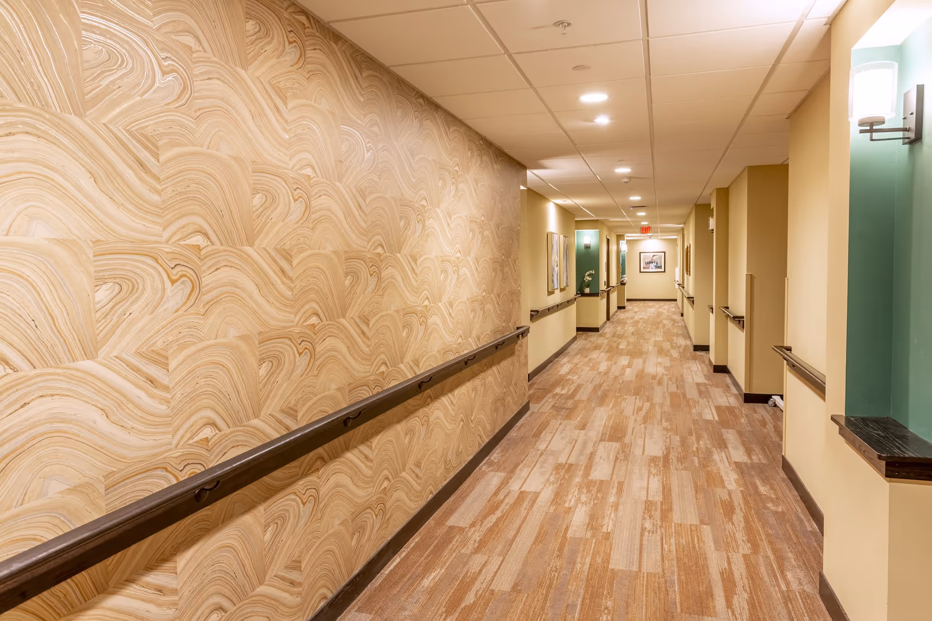 A well-lit indoor hallway with wood-patterned flooring and beige walls. One side of the hallway features a decorative wall with a swirling wood grain pattern, while the other side has beige walls with green accents and mounted handrails. The ceiling has recessed lighting and there are framed pictures hanging along the hallway.