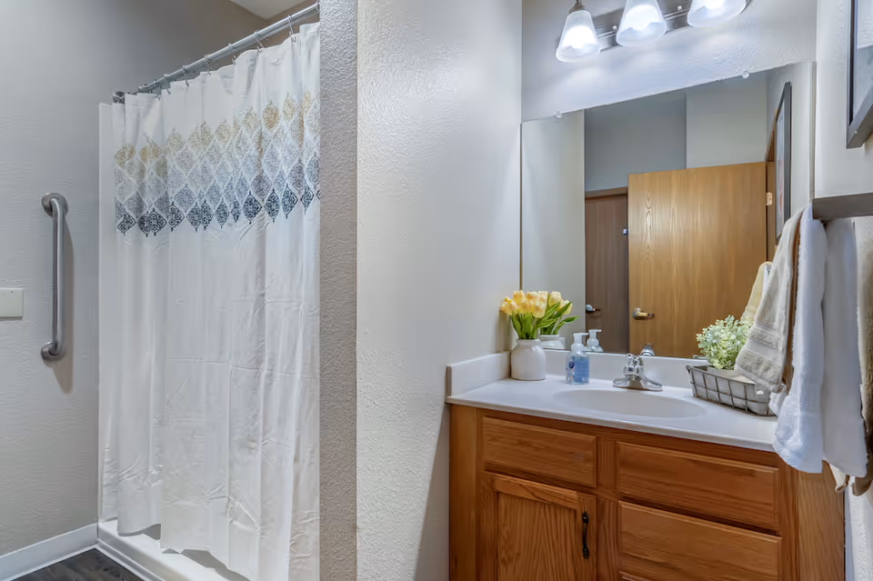 A bathroom with a shower area featuring a white curtain with a decorative pattern, a grab bar on the wall, and a wooden vanity with a white countertop. On the countertop, there is a vase with yellow tulips, a soap dispenser, and a small basket with towels and a plant. A large mirror and three light fixtures are mounted above the sink.