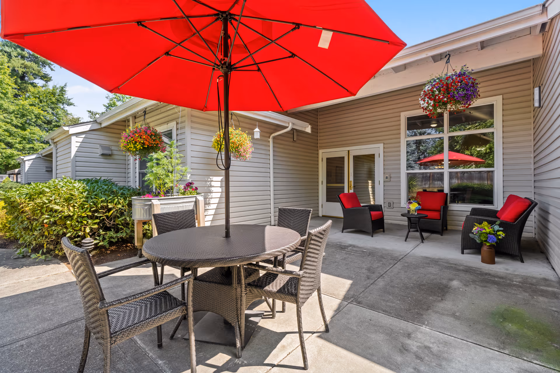 Outdoor patio area with a round table and four wicker chairs under a large red umbrella. There are hanging flower baskets and additional wicker chairs with red cushions near a building with beige siding and large windows.