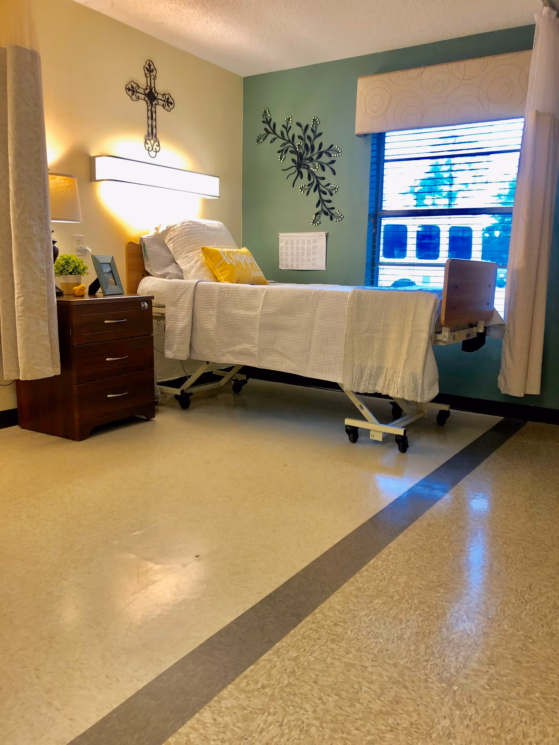 A clean and well-lit patient room in a healthcare facility featuring a hospital bed with white bedding and a yellow pillow, a wooden bedside table with a lamp, a small plant, and a picture frame. The walls are painted in light green and beige, decorated with a metal cross and a floral wall art. A window with blinds and curtains lets in natural light.