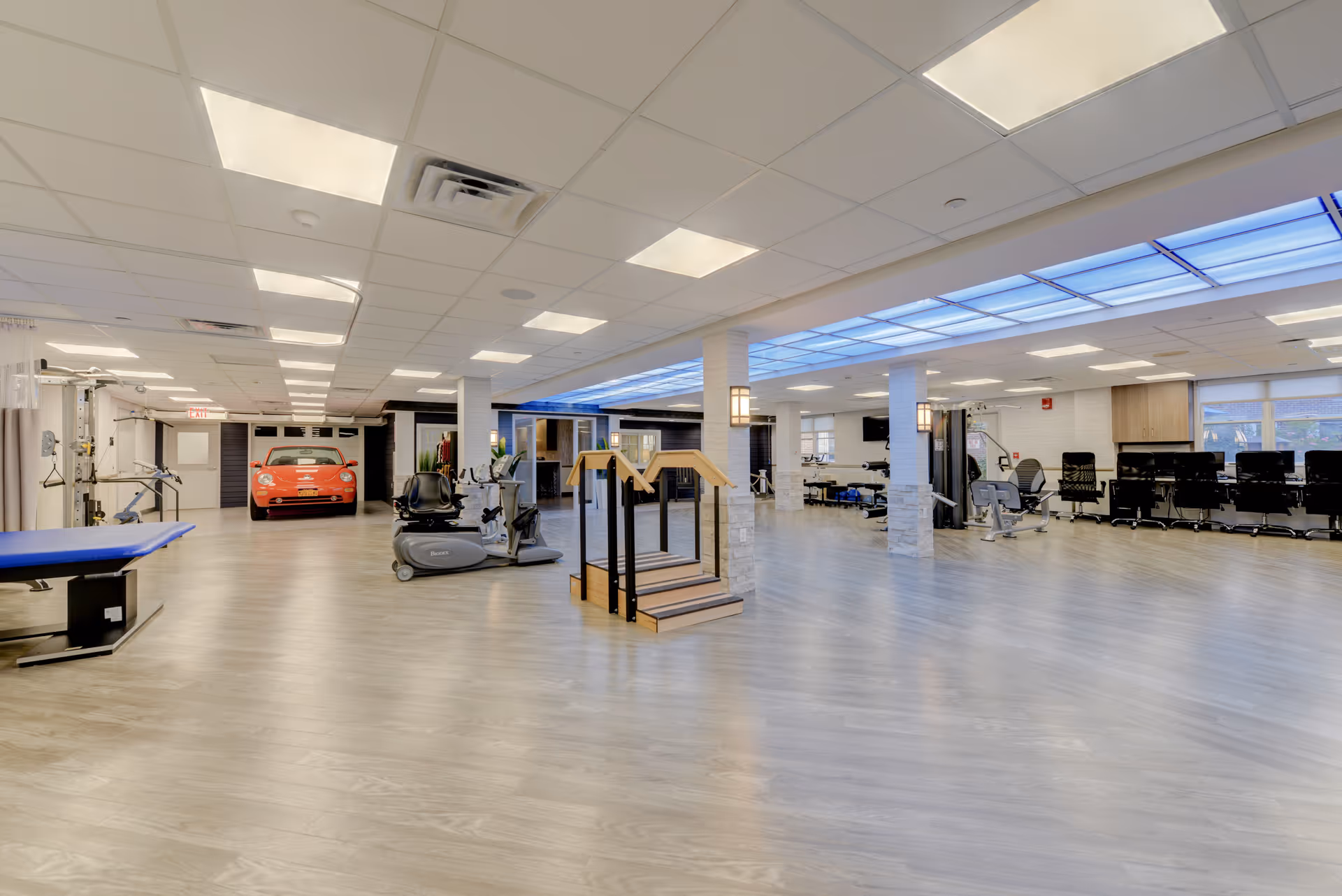 A spacious rehabilitation and fitness room with exercise equipment including a recumbent bike, parallel bars for walking practice, and various other physical therapy machines. The room has a light wood floor, white ceiling with recessed lighting, and a blue-lit ceiling panel. There is a red car positioned at the far end near an exit door, and a row of black chairs and a TV mounted on the wall to the right.