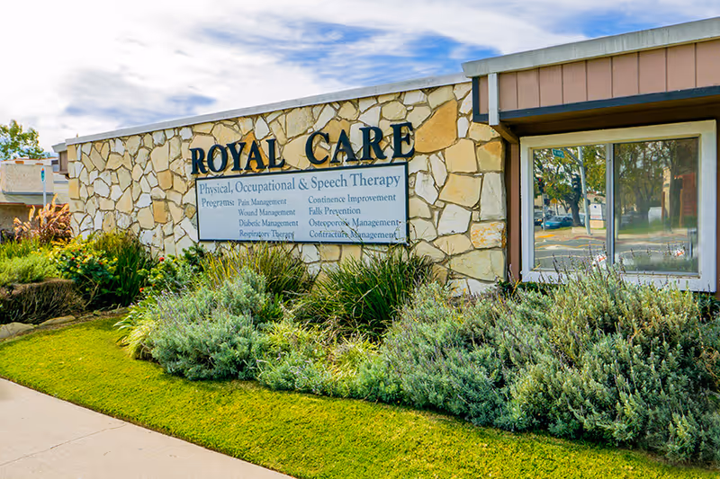 Exterior view of Royal Care Skilled Nursing Center showing a stone wall with the facility's name and a sign listing physical, occupational, and speech therapy programs offered, surrounded by green shrubs and grass under a partly cloudy sky.