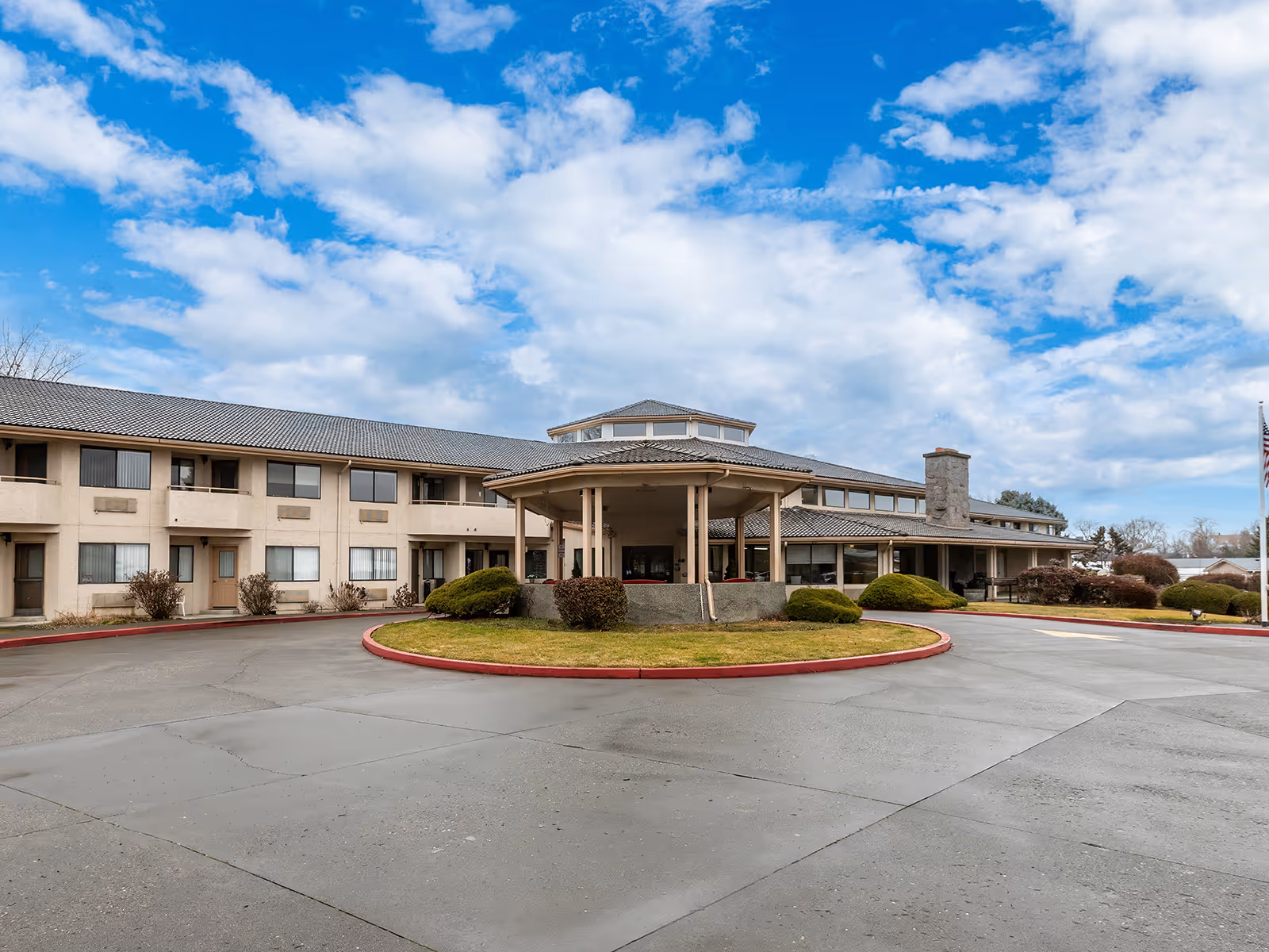 Exterior view of a two-story senior living facility building with a covered entrance, surrounded by a circular driveway and landscaped bushes under a partly cloudy blue sky.