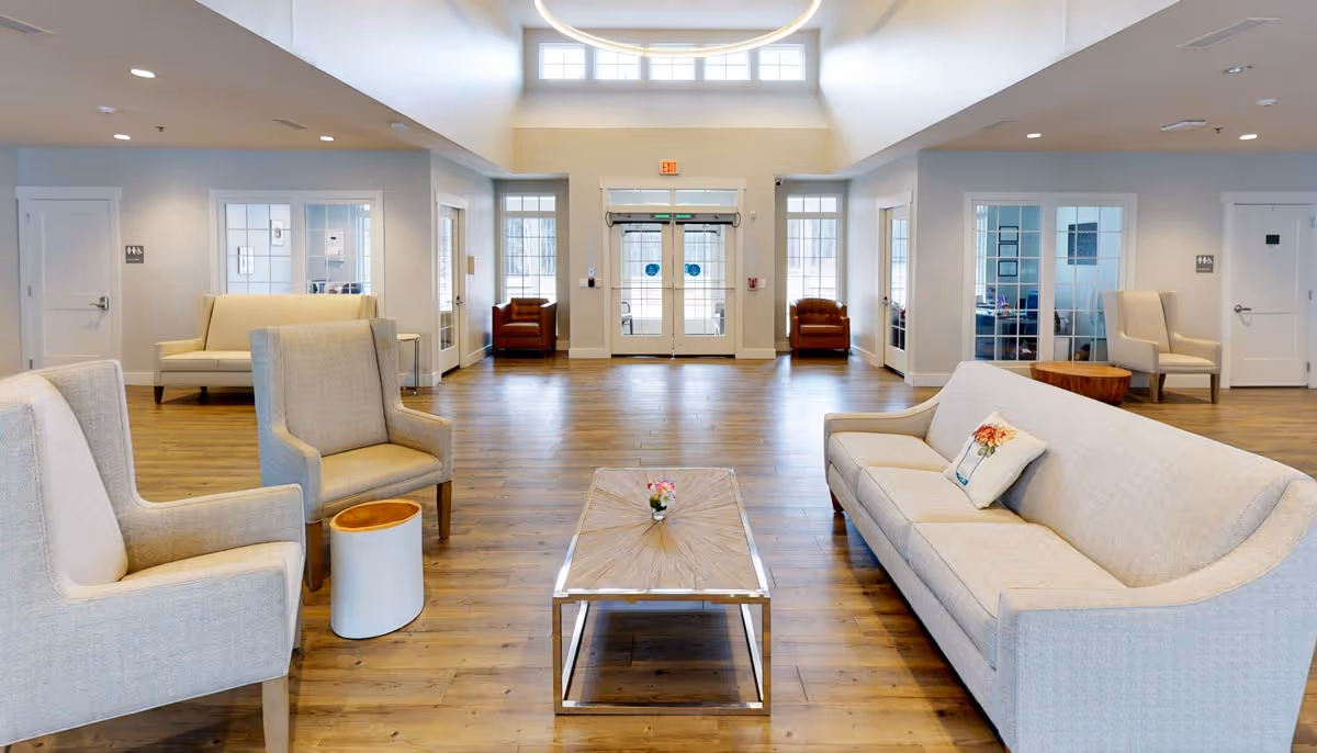 A spacious and well-lit living room area with light-colored sofas and armchairs arranged around a wooden coffee table with a small flower vase. The room has large windows and glass doors letting in natural light, wooden flooring, and neutral-colored walls. There are additional seating areas and doors leading to other rooms visible in the background.