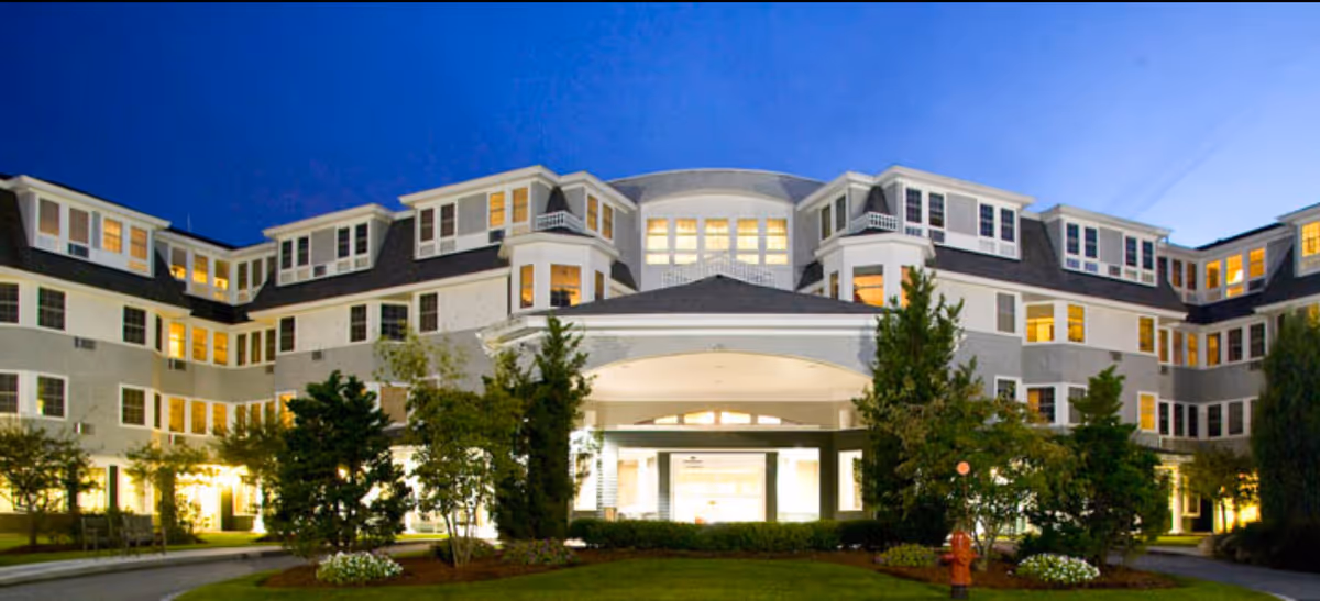 Exterior view of a large, well-lit senior living facility building at dusk with multiple windows and a covered entrance surrounded by landscaped greenery and trees.