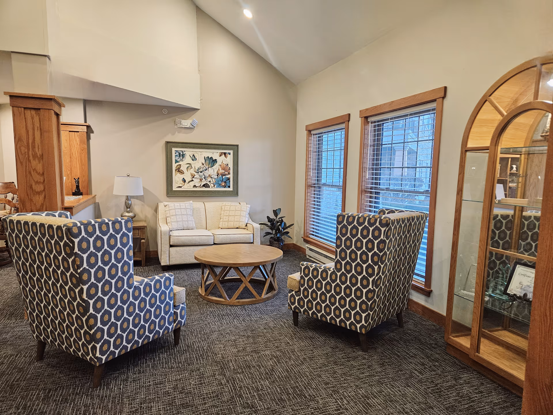 A cozy senior living common room with a sofa, two patterned armchairs, a round coffee table, and a display cabinet.