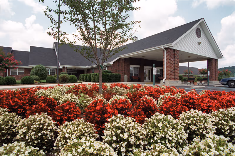 Front exterior view of Lynchburg Nursing Center showing a brick building with a covered entrance, surrounded by well-maintained landscaping including red and white flowers and green shrubs under a partly cloudy sky.